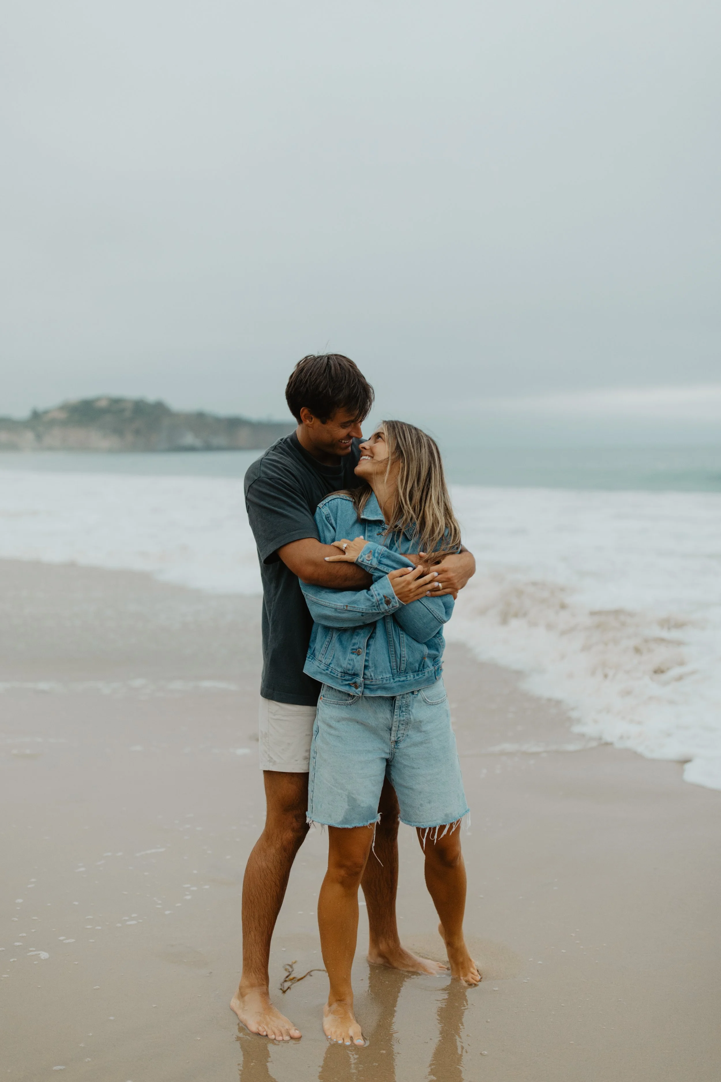 A couple embracing on a beach during cloudy weather, with the ocean and distant cliffs in the background.
