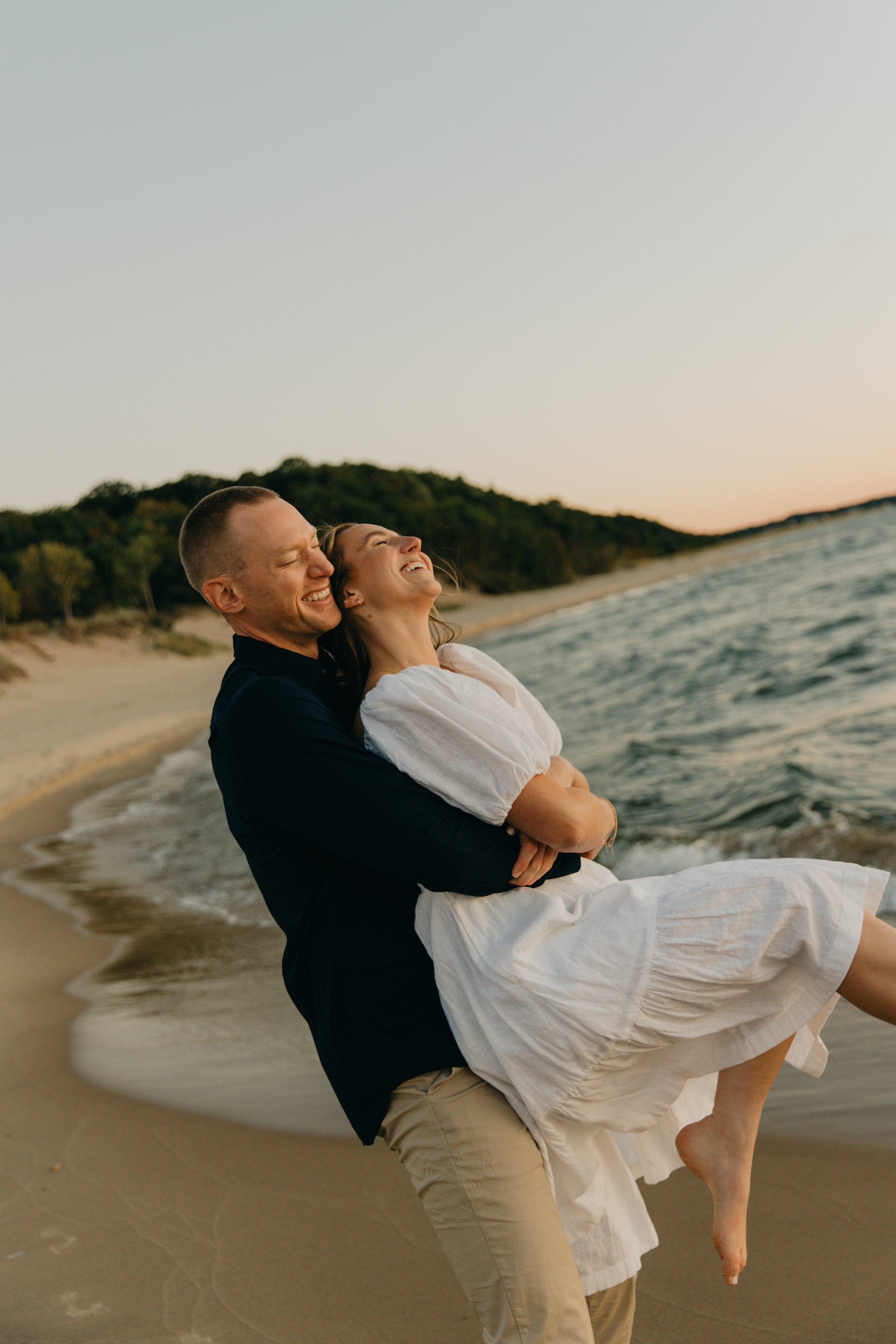 A couple laughing and embracing on a beach during sunset.