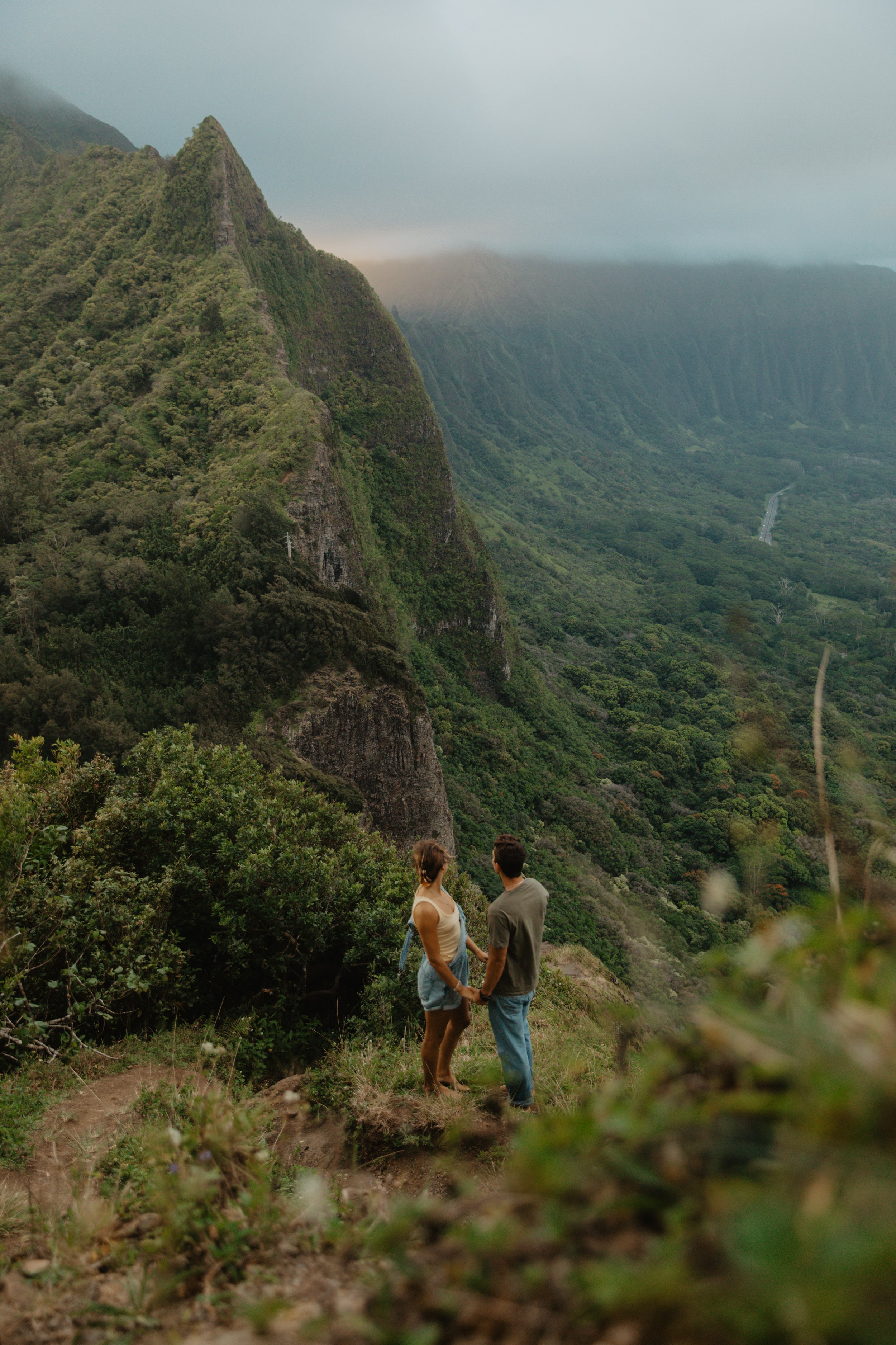 A couple holding hands and looking at a lush green mountain landscape with steep cliffs and dense forest.