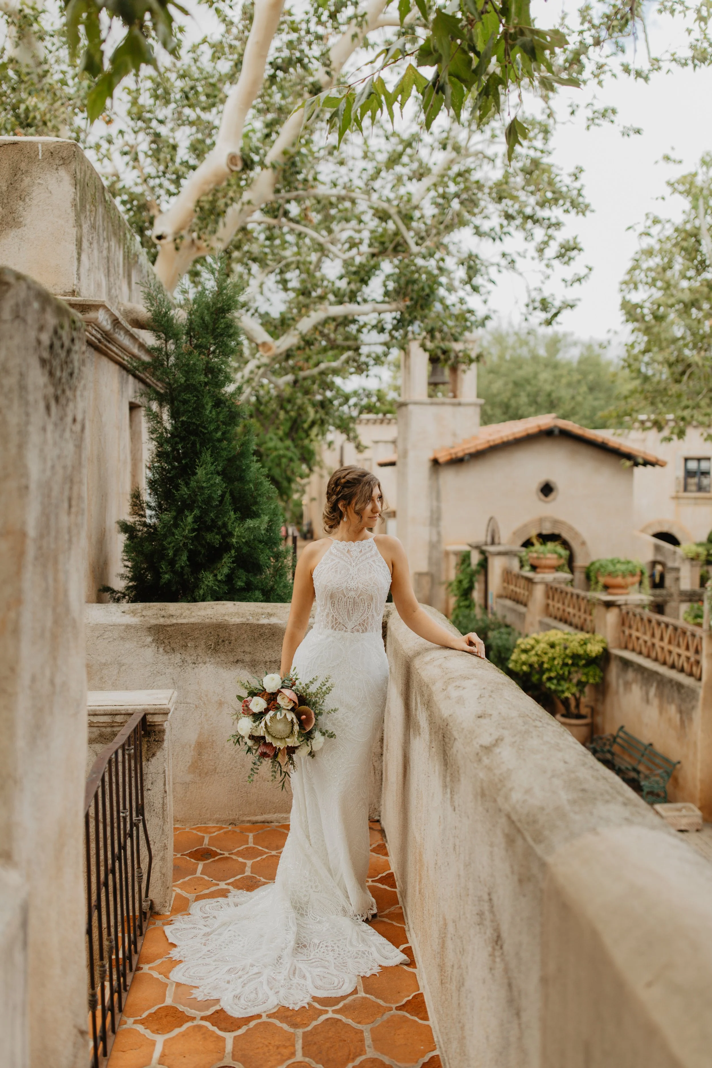 A bride in a white lace wedding dress holding a floral bouquet stands on a stone balcony with a rustic Mediterranean-style building and greenery in the background.