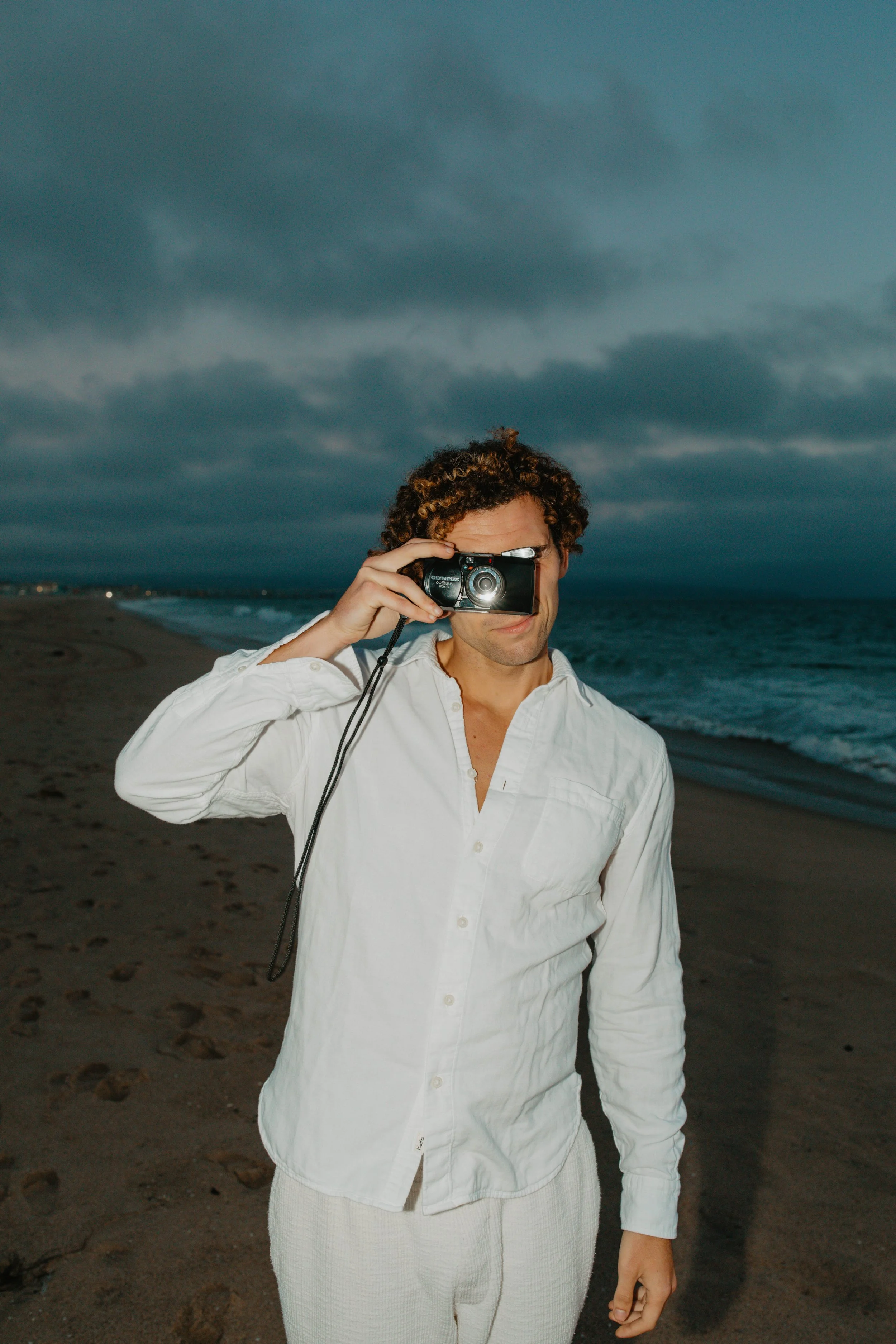 A man with curly hair taking a photo with a camera on a beach in cloudy weather.