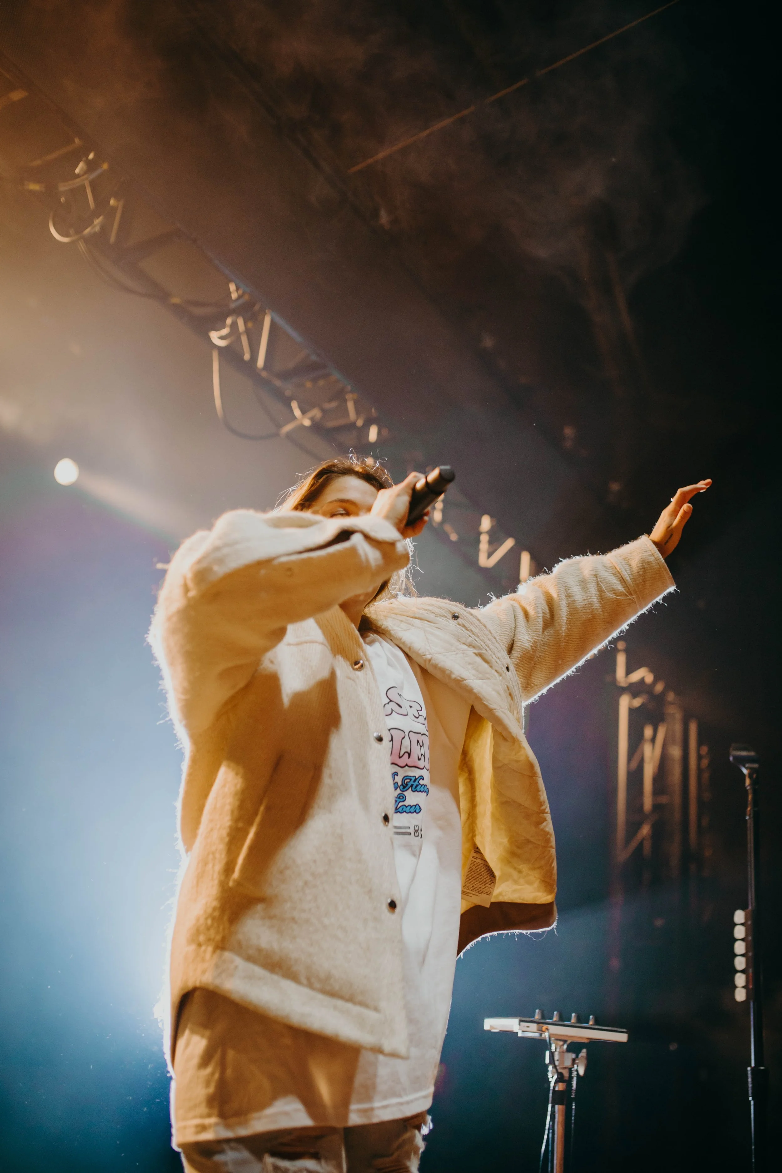 Chelea Cutler performing on stage, holding a microphone and raising her hand, with stage lights and equipment in the background.