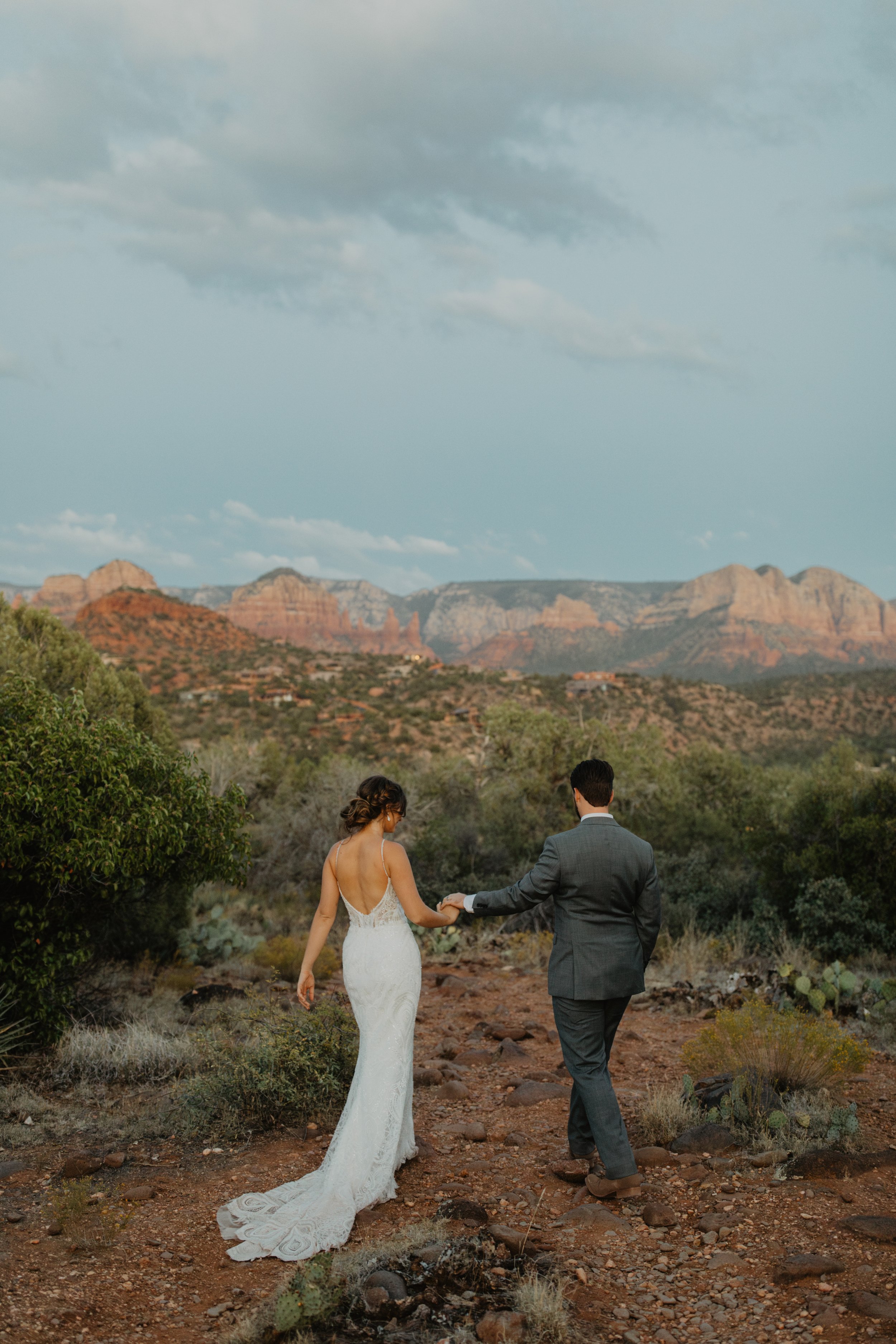 A bride and groom walking hand in hand through a desert landscape with rocky hills and mountains in the background.