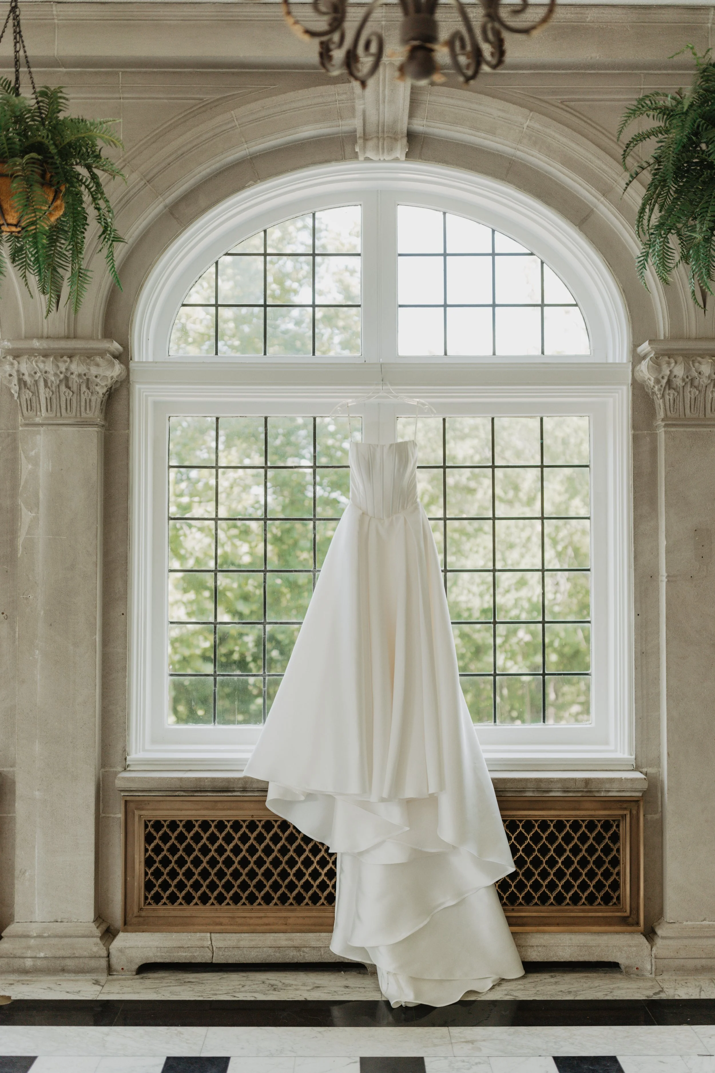 A white wedding dress hanging in front of a large arched window with a grid pattern, surrounded by stone columns and decorated with hanging ferns.