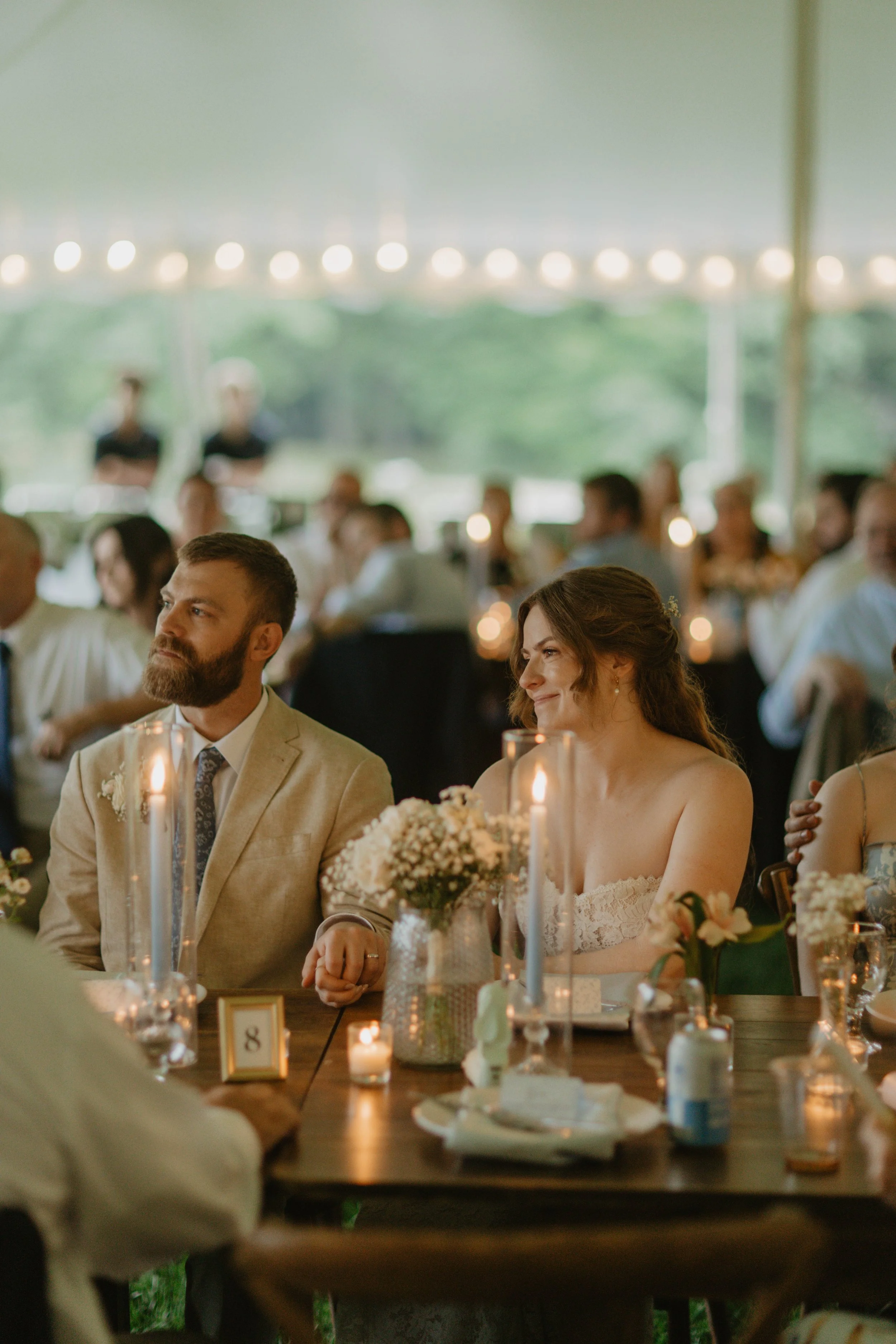 Bride and groom sitting at a wedding reception table decorated with candles and flowers during their reception, with guests and string lights in the background.