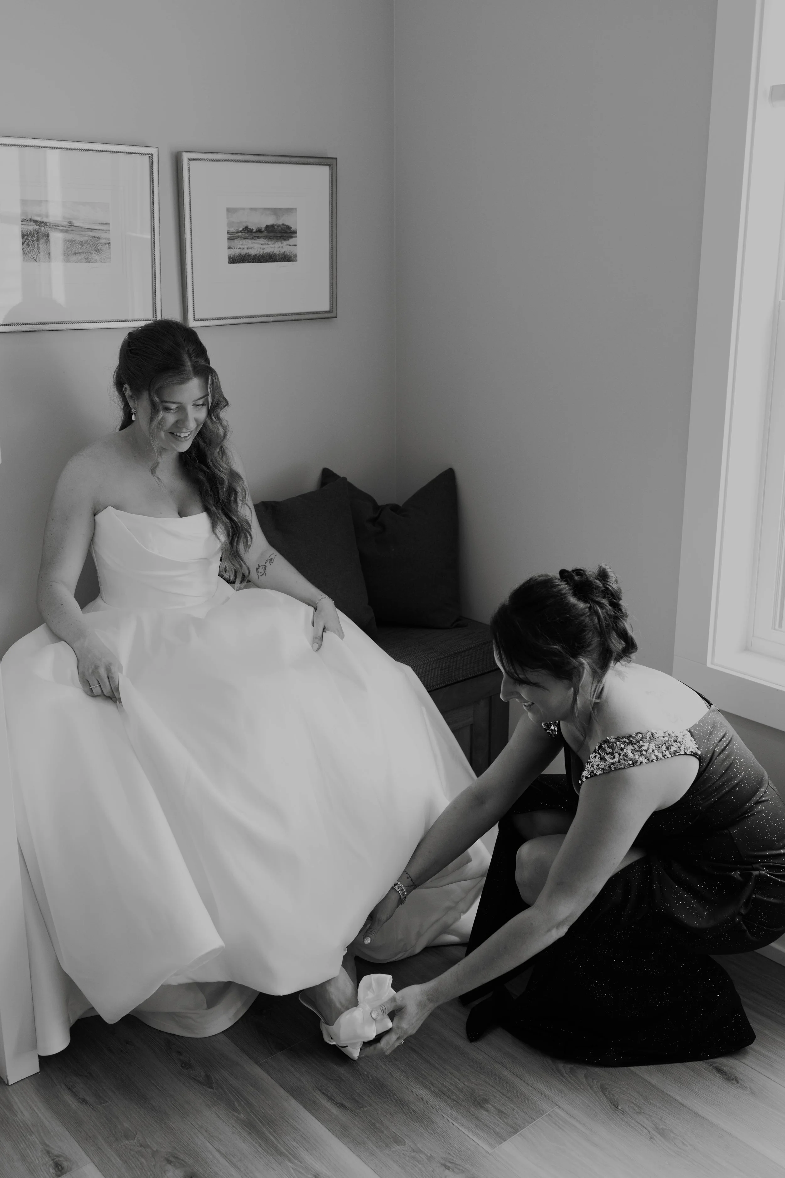 A bride in a white wedding gown sits on a bench, smiling, as a woman helps her put on her shoe in a well-lit room with framed pictures on the wall.