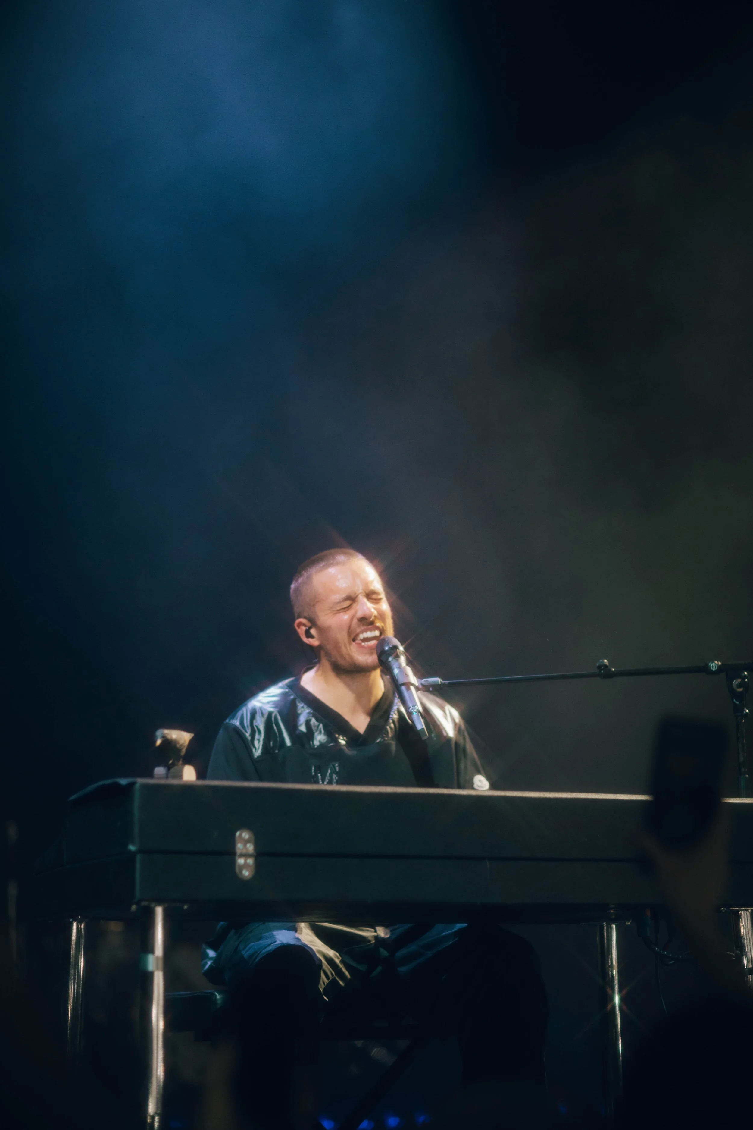 A man passionately singing and playing the keyboard on stage with a microphone in front of him.