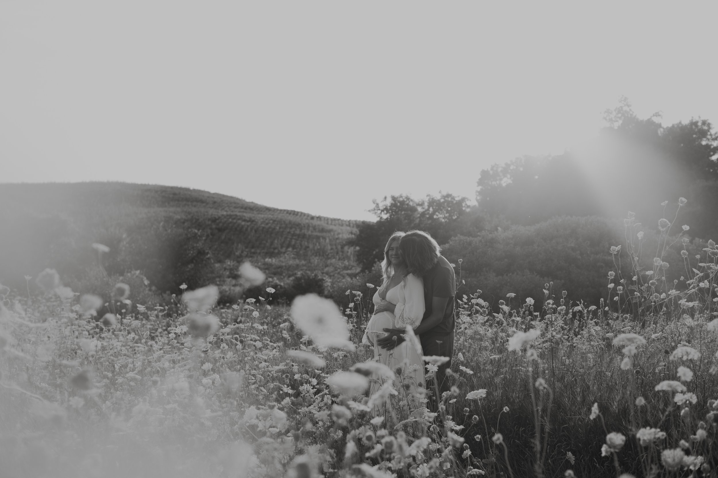A black and white photo of a couple standing in a field of flowers, with the woman cradling her pregnant belly, and a scenic landscape in the background.