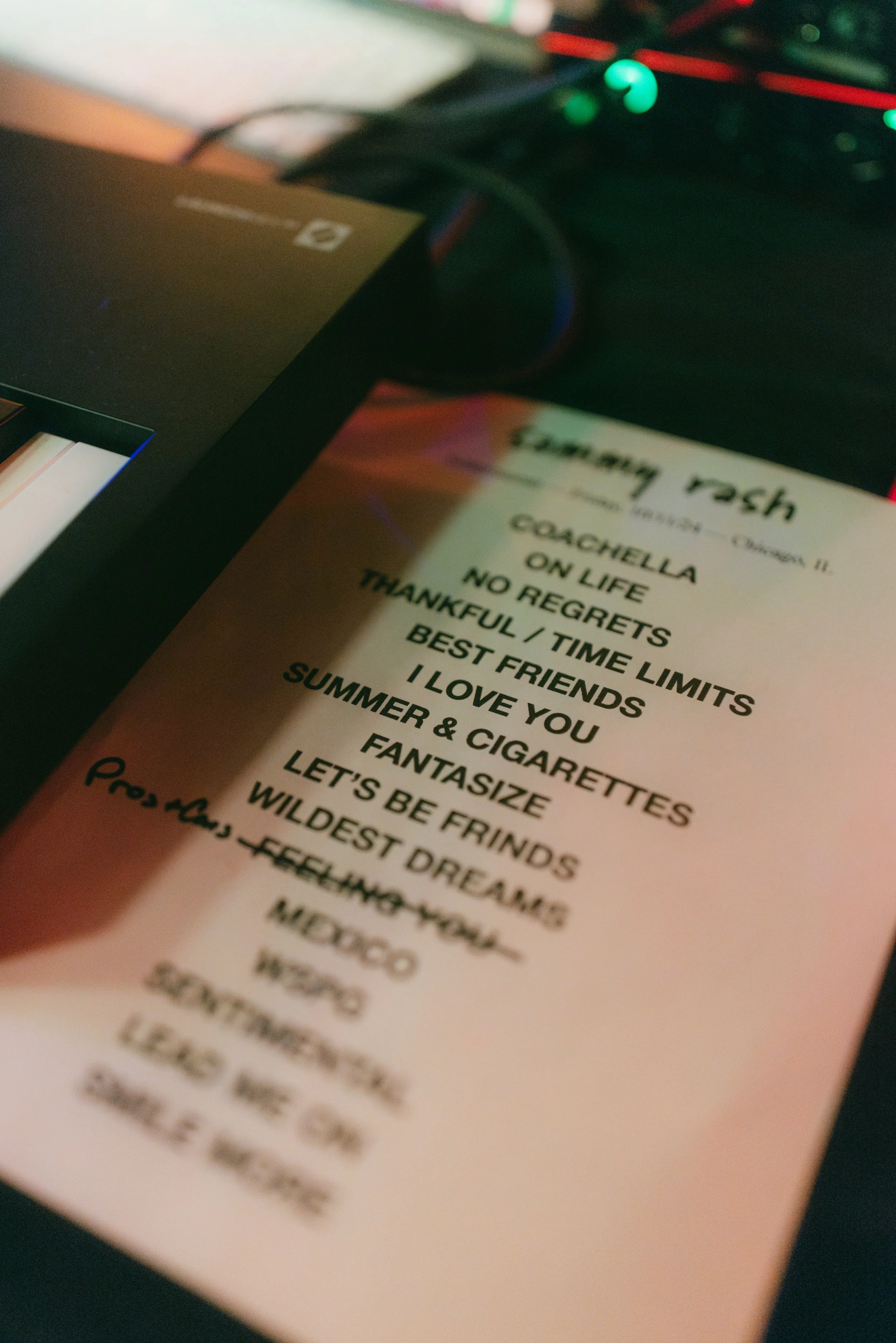 Close-up of a printed song list titled 'Sammy Rash'' on a piece of paper, with some headlines like 'Cochella,' 'On Life,' 'No Regrets,' 'Thankful / Time Limits,' and others, placed on a desk with electronic equipment.