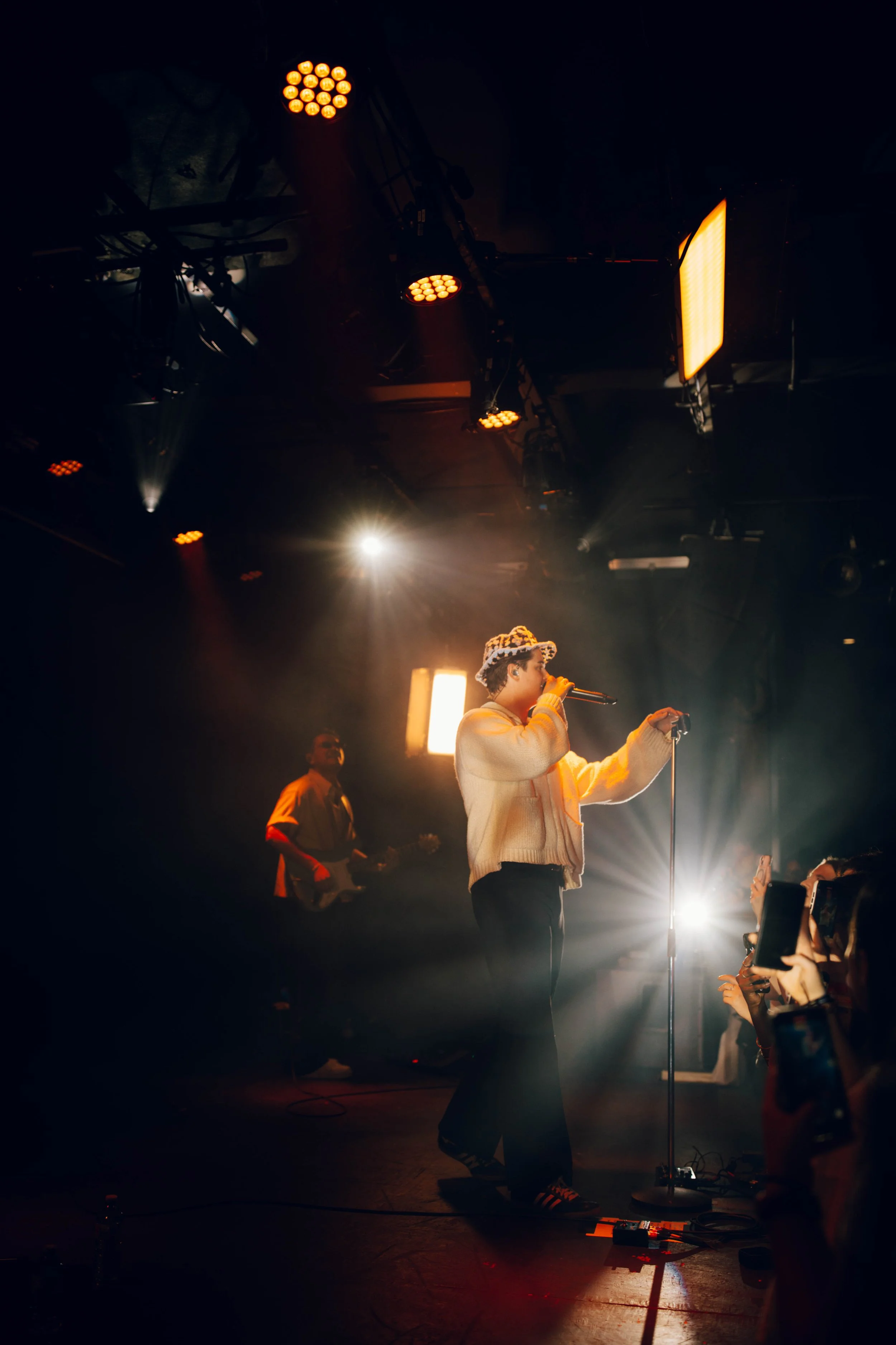 The music artist, Nicky Youre performing on stage, holding a microphone and singing. The stage is lit with spotlights and stage lights.