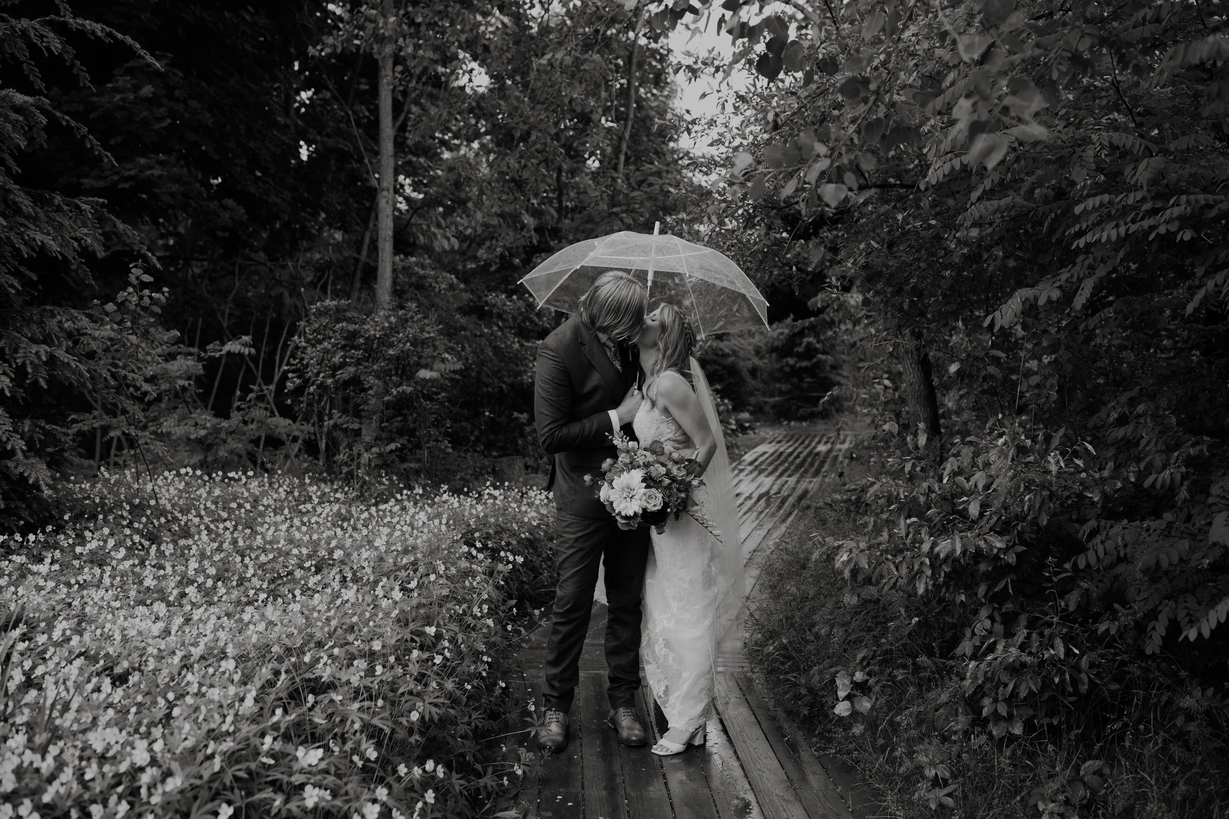 A black and white photo of a couple kissing under an umbrella on a wooden path in a garden, with flowers and trees around.