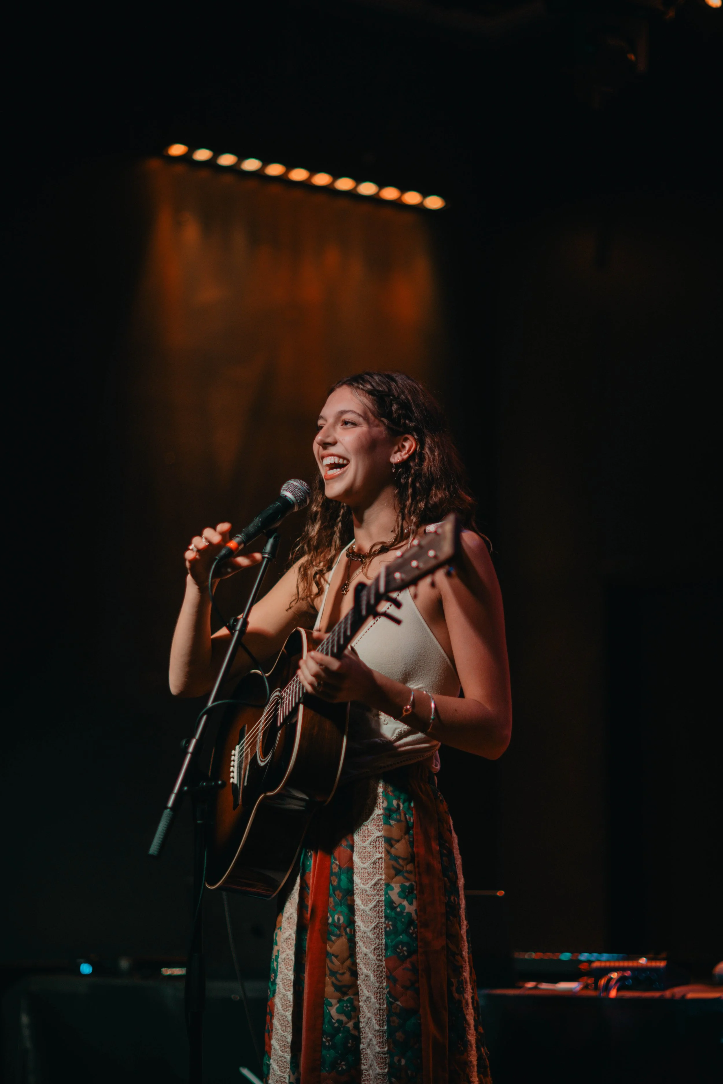 The music artist Emma Andersen singing into a microphone while playing an acoustic guitar on stage, smiling cheerfully, with warm lighting in the background.