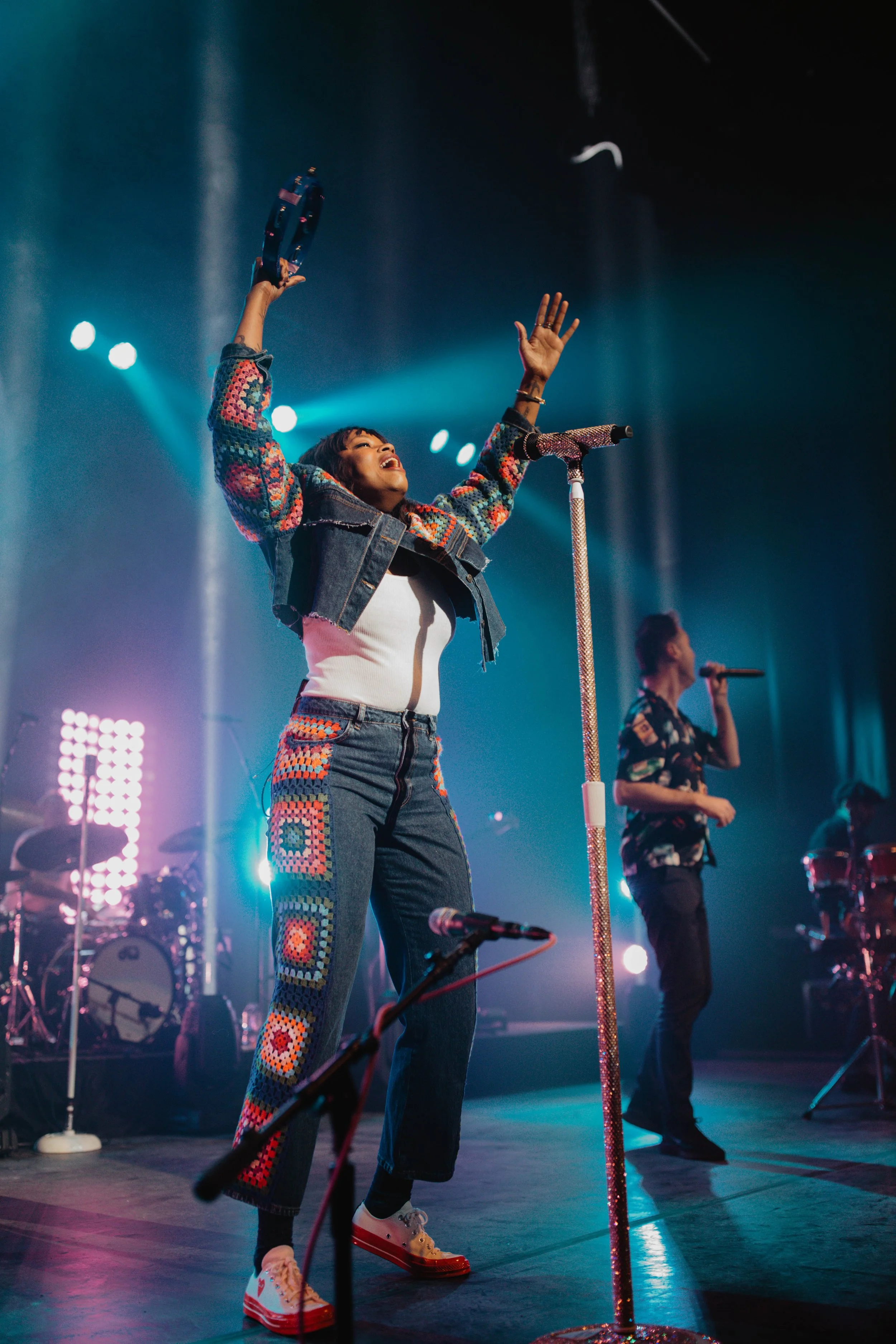 The band Fitz and the Tantrams performing with colorful crochet jacket and jeans dancing on stage with her arms raised, accompanied by a musician singing into a microphone, with stage lights and drums in the background in Chicago.