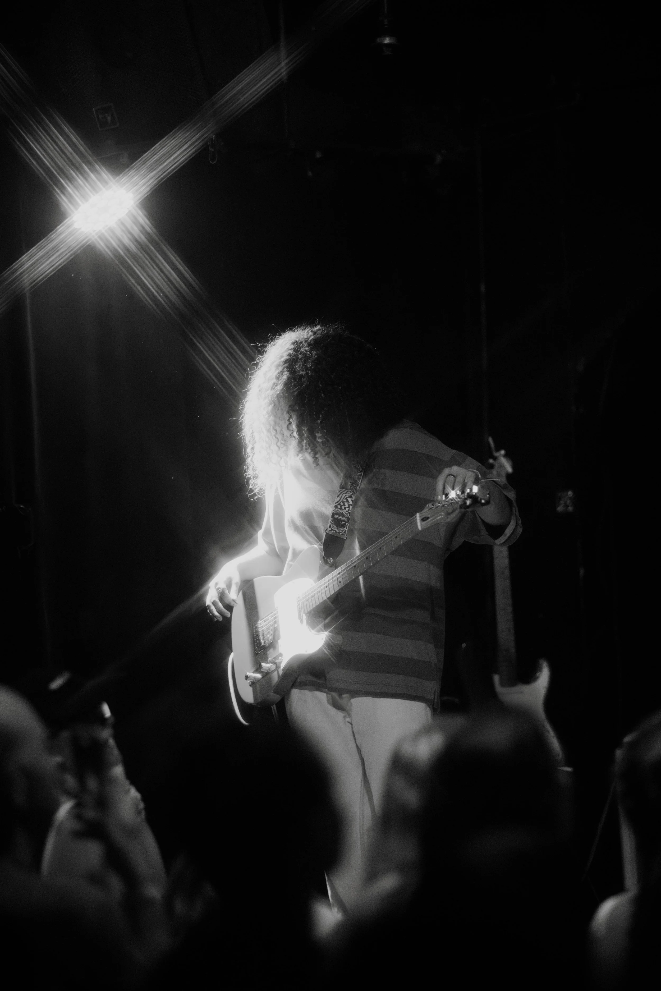 A person with curly hair playing an electric guitar on stage, backlit by bright lighting, with an audience in the foreground.