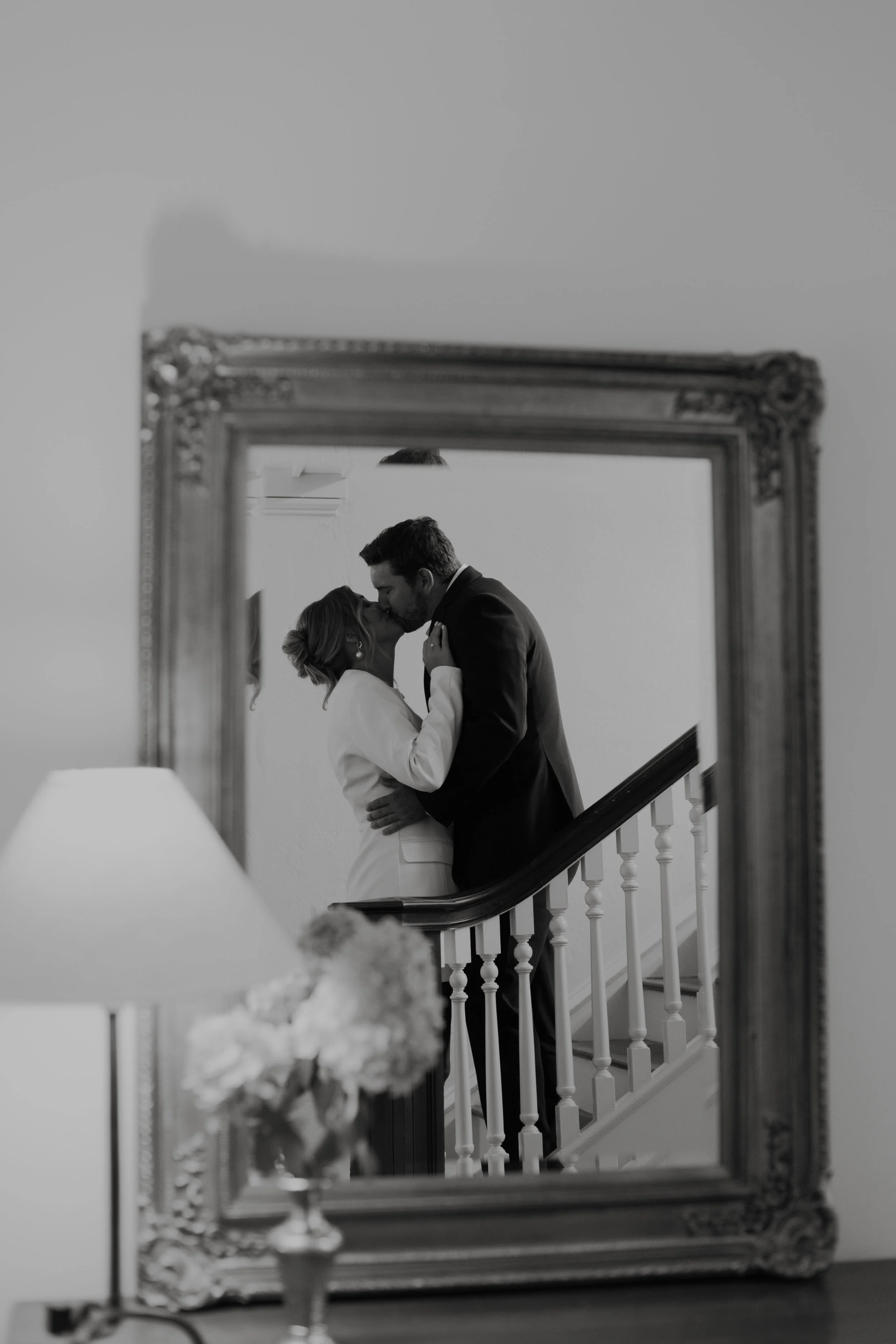 Black and white photo of a couple kissing on a staircase, seen through an ornate mirror, with a lamp and flowers in the foreground.