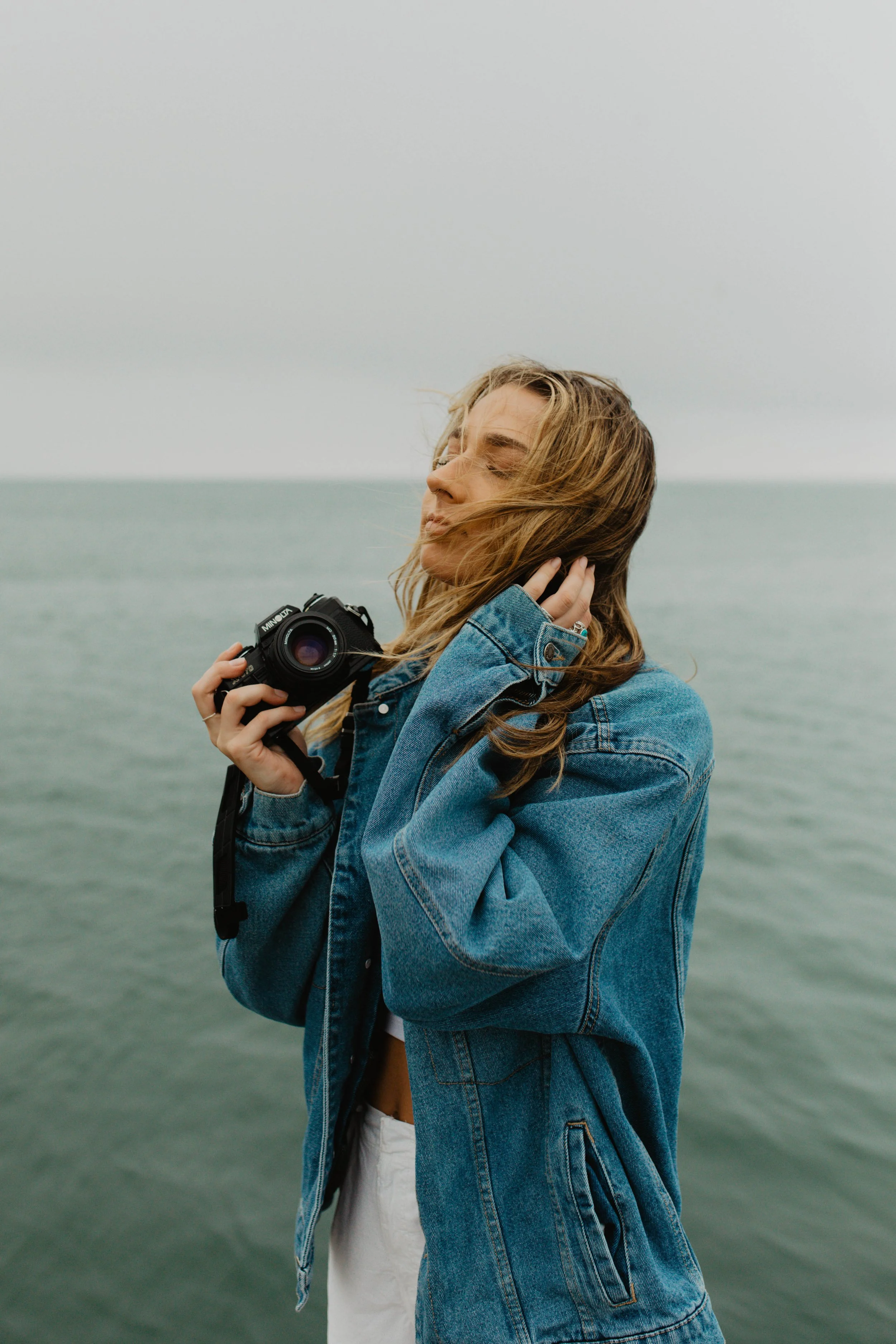 A woman holding a camera with one hand and gently touching her hair with the other, standing near water, eyes closed, hair blowing in the wind, wearing a denim jacket.