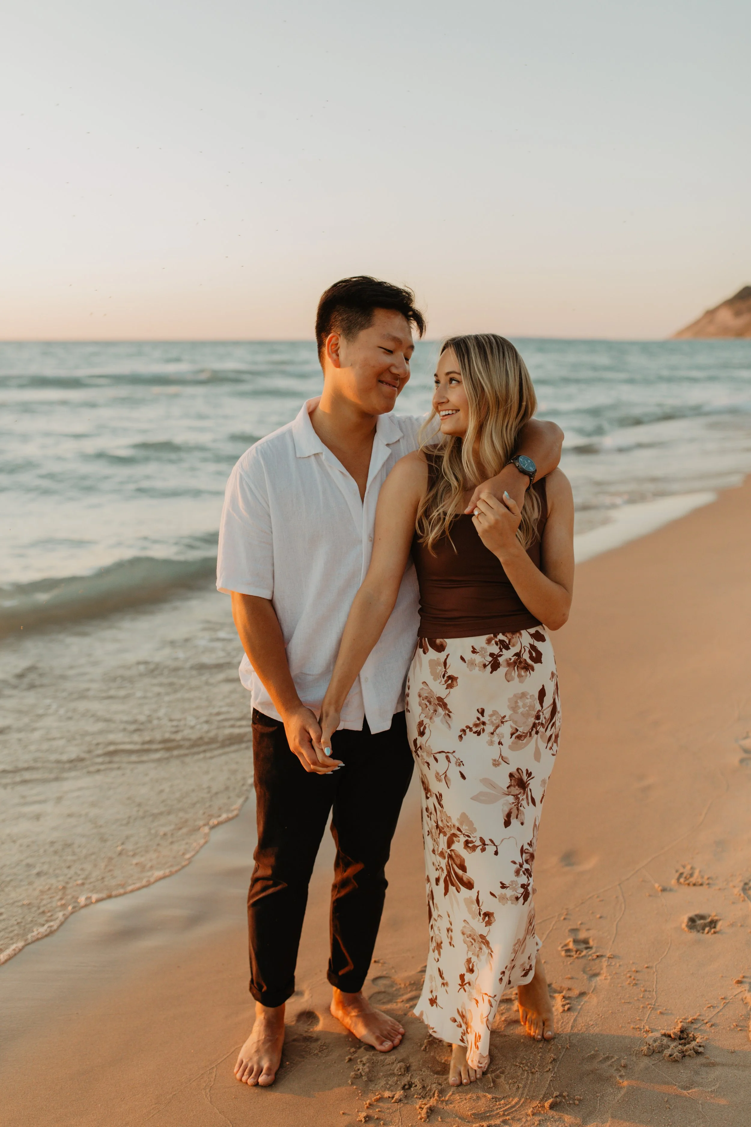 A happy couple walking barefoot on the beach during sunset, holding hands and smiling at each other.