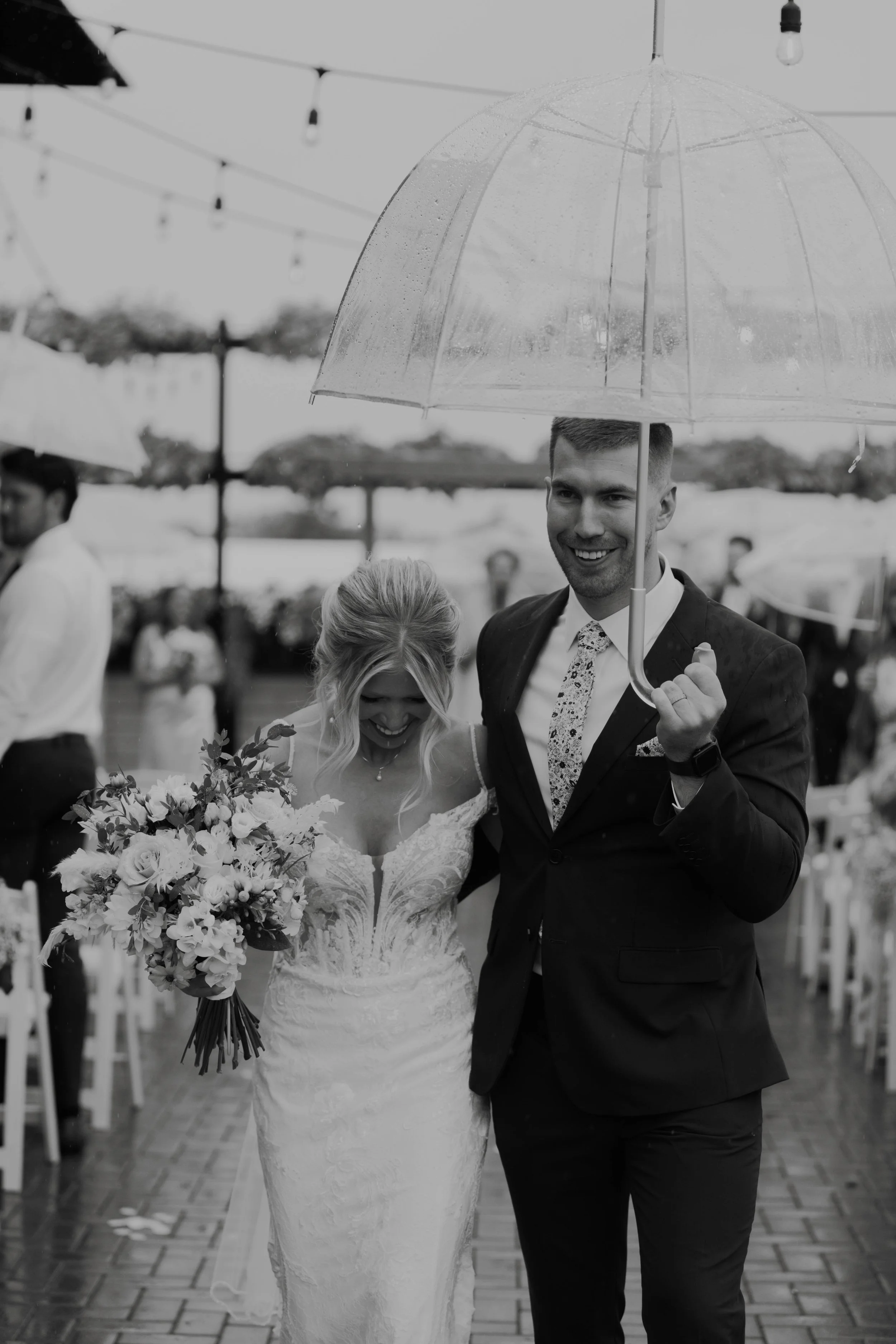 A bride and groom walking together under an umbrella at their wedding, smiling and holding a bouquet of flowers.