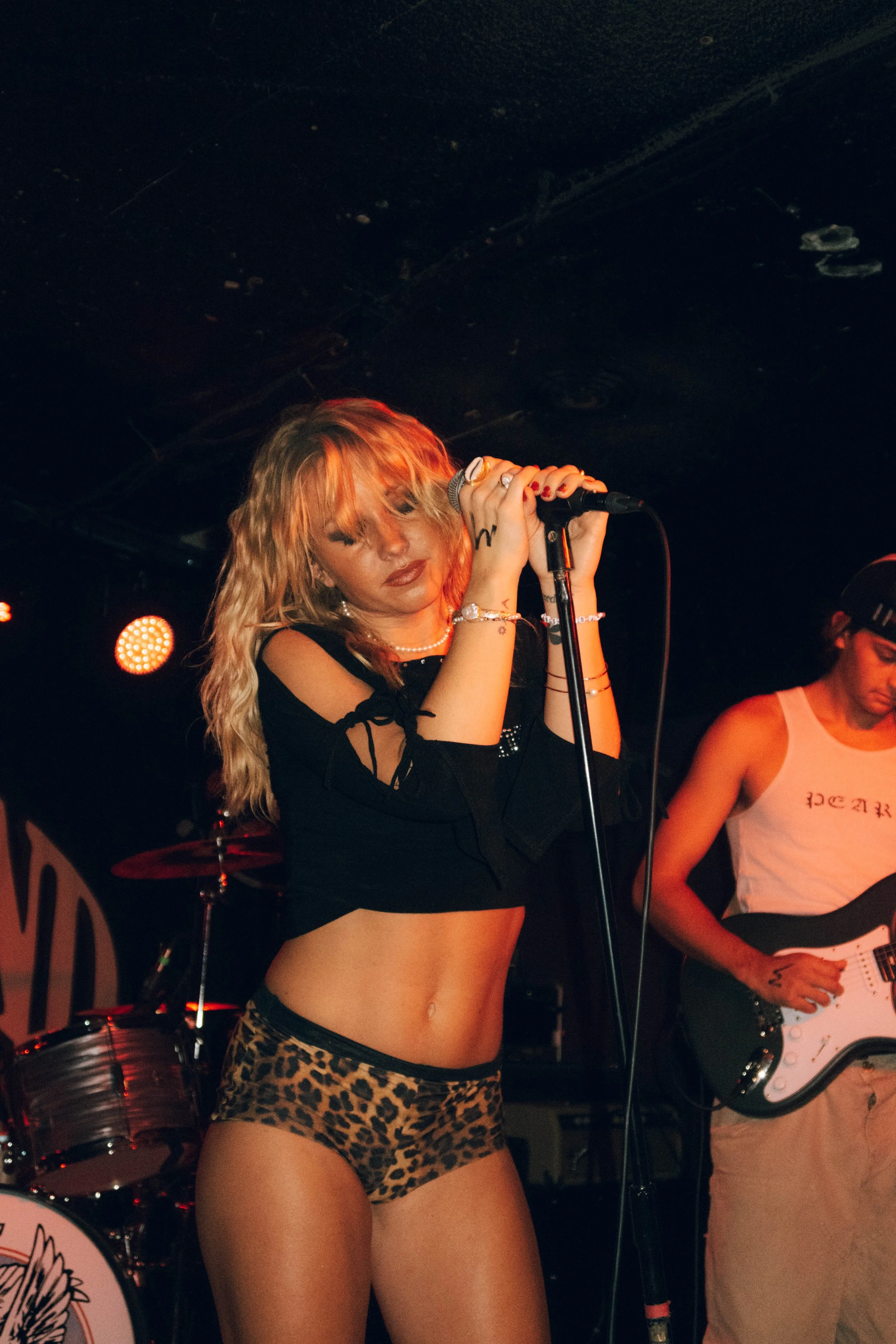 The music artist PEARL singing into a microphone on stage, wearing a black crop top with cut-out shoulders and leopard-print shorts, with a man playing an electric guitar in the background at a concert in Detroit, Michigan.