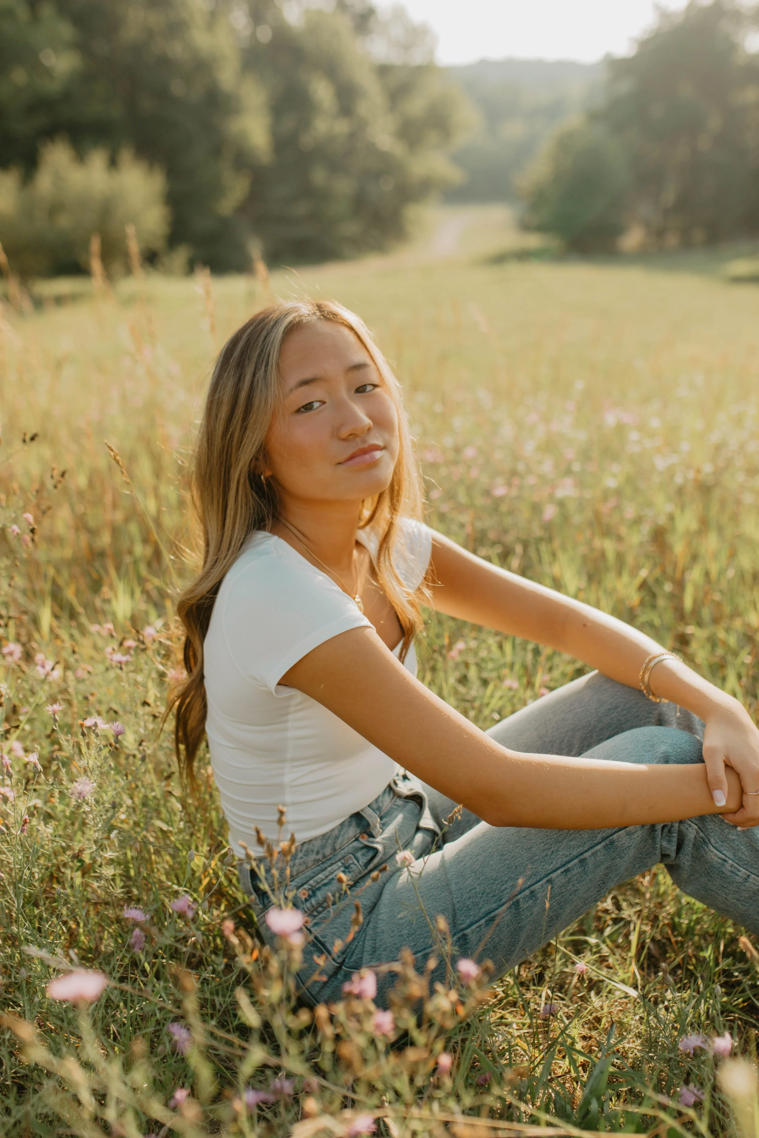 Young woman sitting in a field of grass and flowers, looking at the camera, with trees and a dirt path in the background.