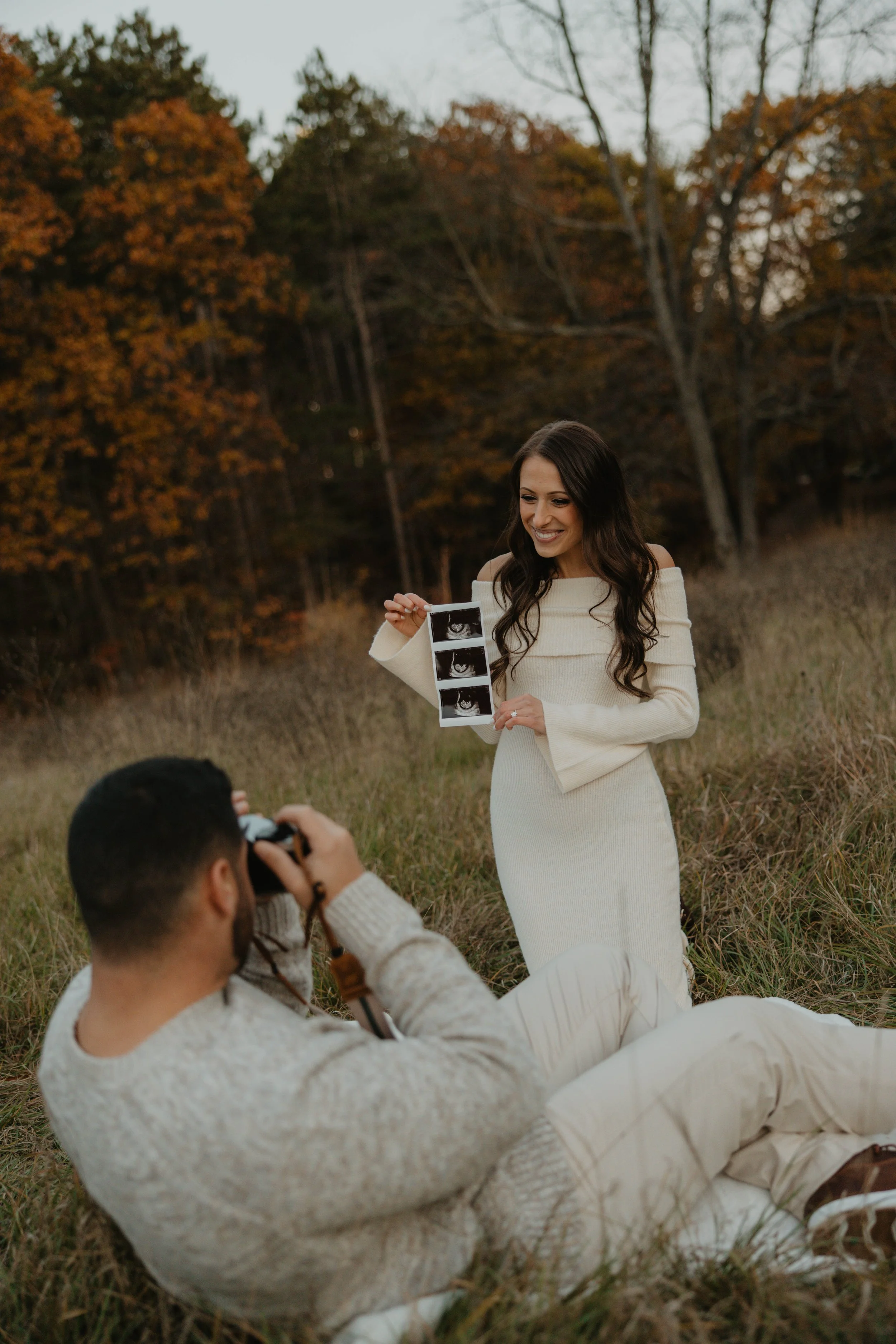 A woman in a white off-the-shoulder dress holds up ultrasound images while a man in a beige sweater and pants takes a photo of her with a camera. They are outdoors in a grassy field with autumn-colored trees in the background.