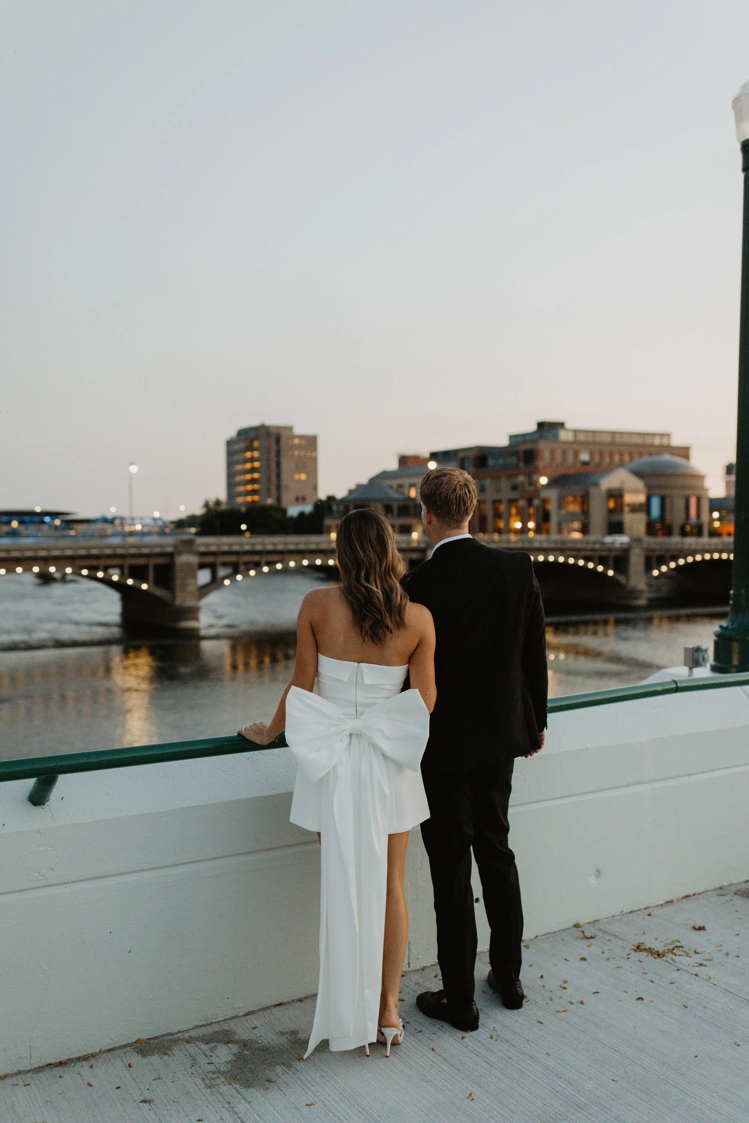 A couple in formal attire, a woman in a white strapless dress with a large bow on the back and high heels, and a man in a black suit, stand on a bridge overlooking a river at sunset, with city buildings in the background.