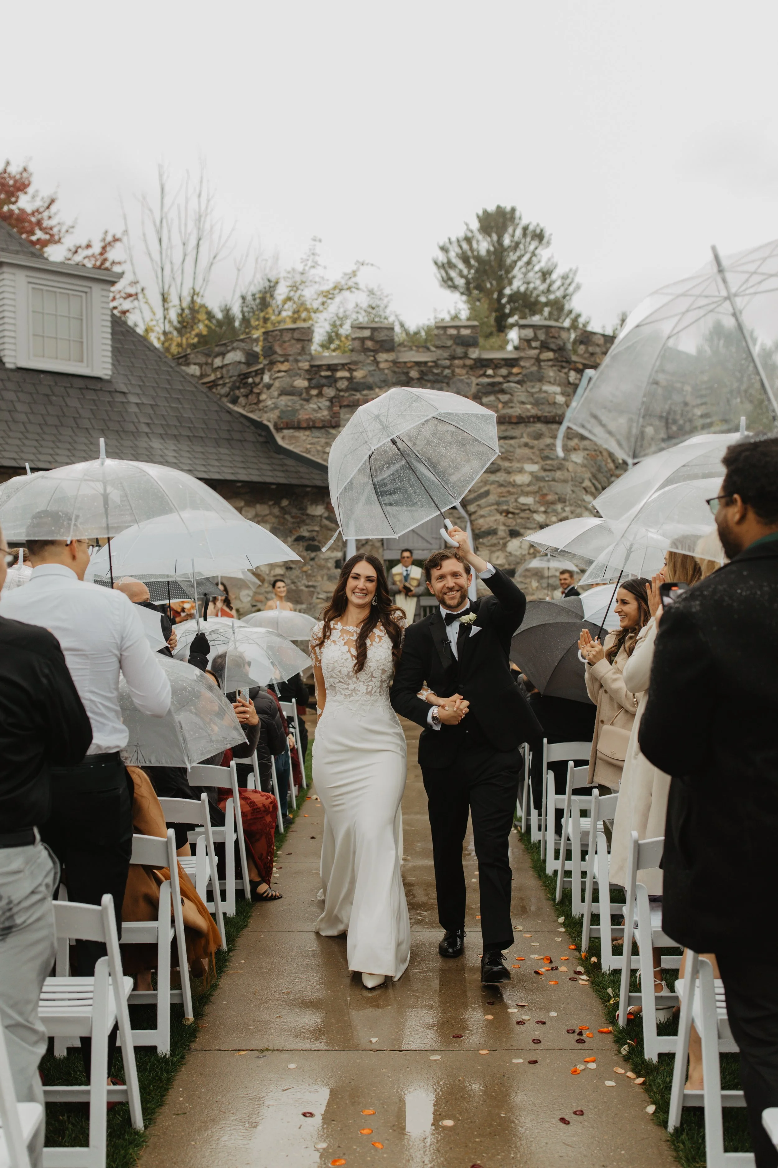 A newlywed couple walking down the aisle at their outdoor wedding ceremony, smiling and holding hands, as guests cheer and applaud under umbrellas on a rainy day.