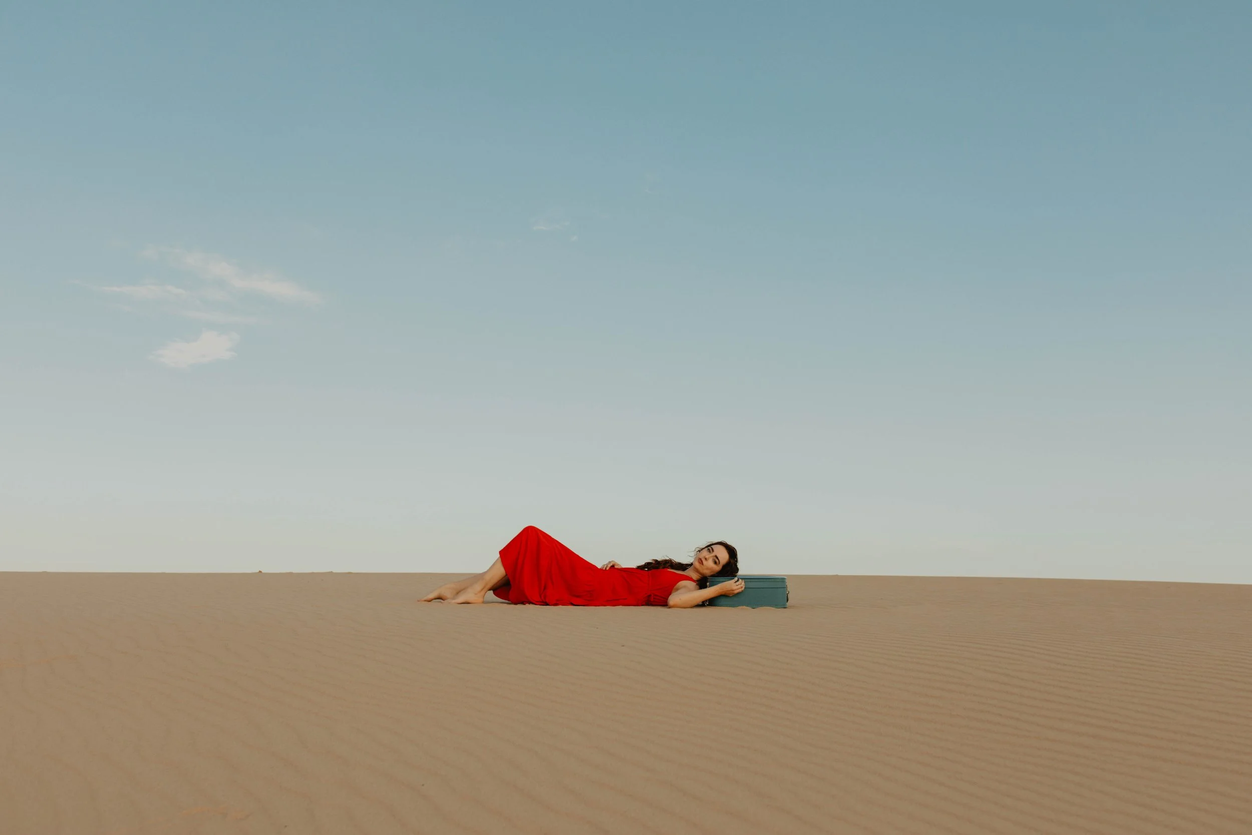 A woman in a red dress lying on sand dunes in a desert, resting her head on a blue suitcase, under a clear sky.