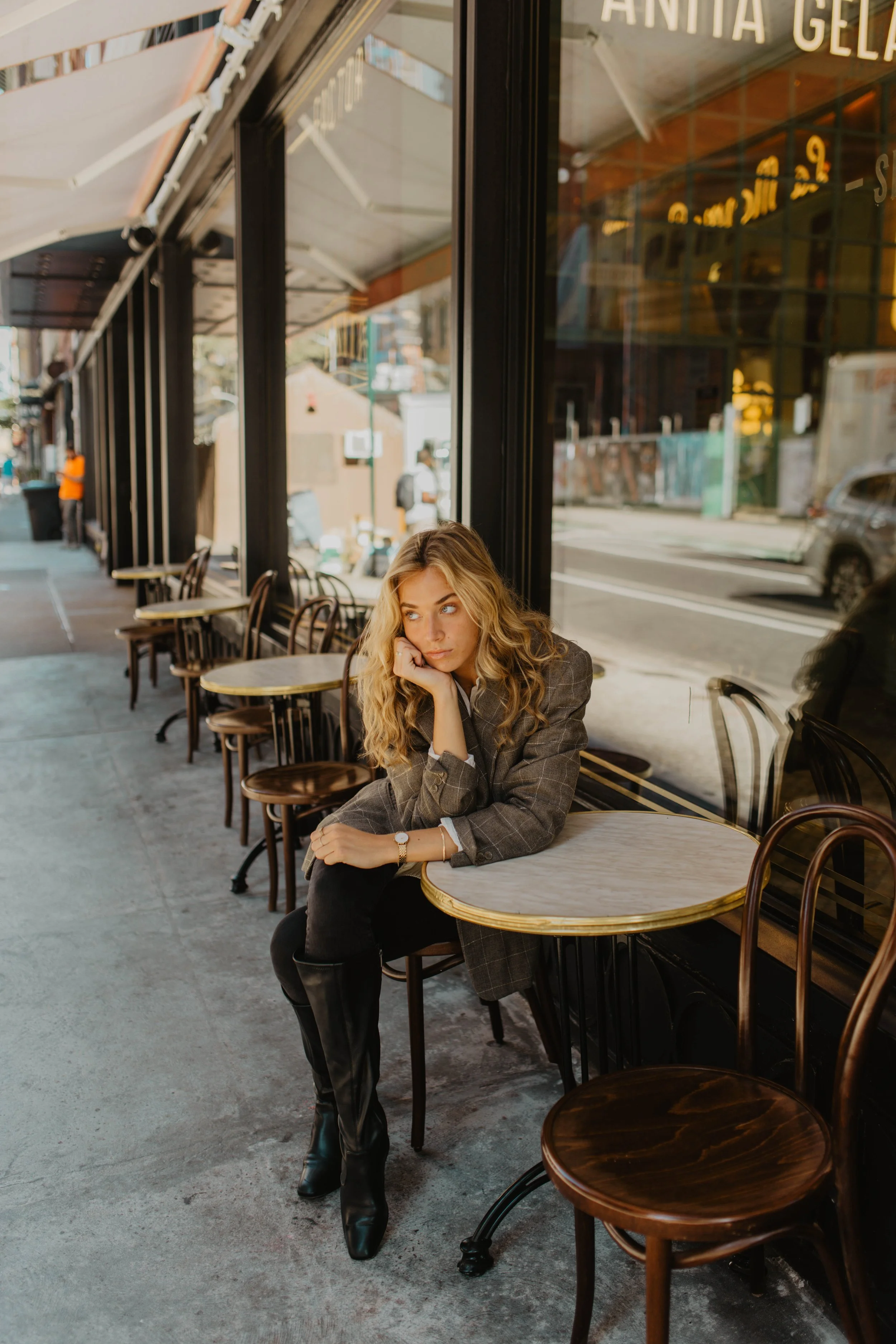 A woman with curly blonde hair sitting alone outside a cafe, resting her chin on her hand, looking thoughtful. She is wearing a plaid blazer, black pants, and boots.