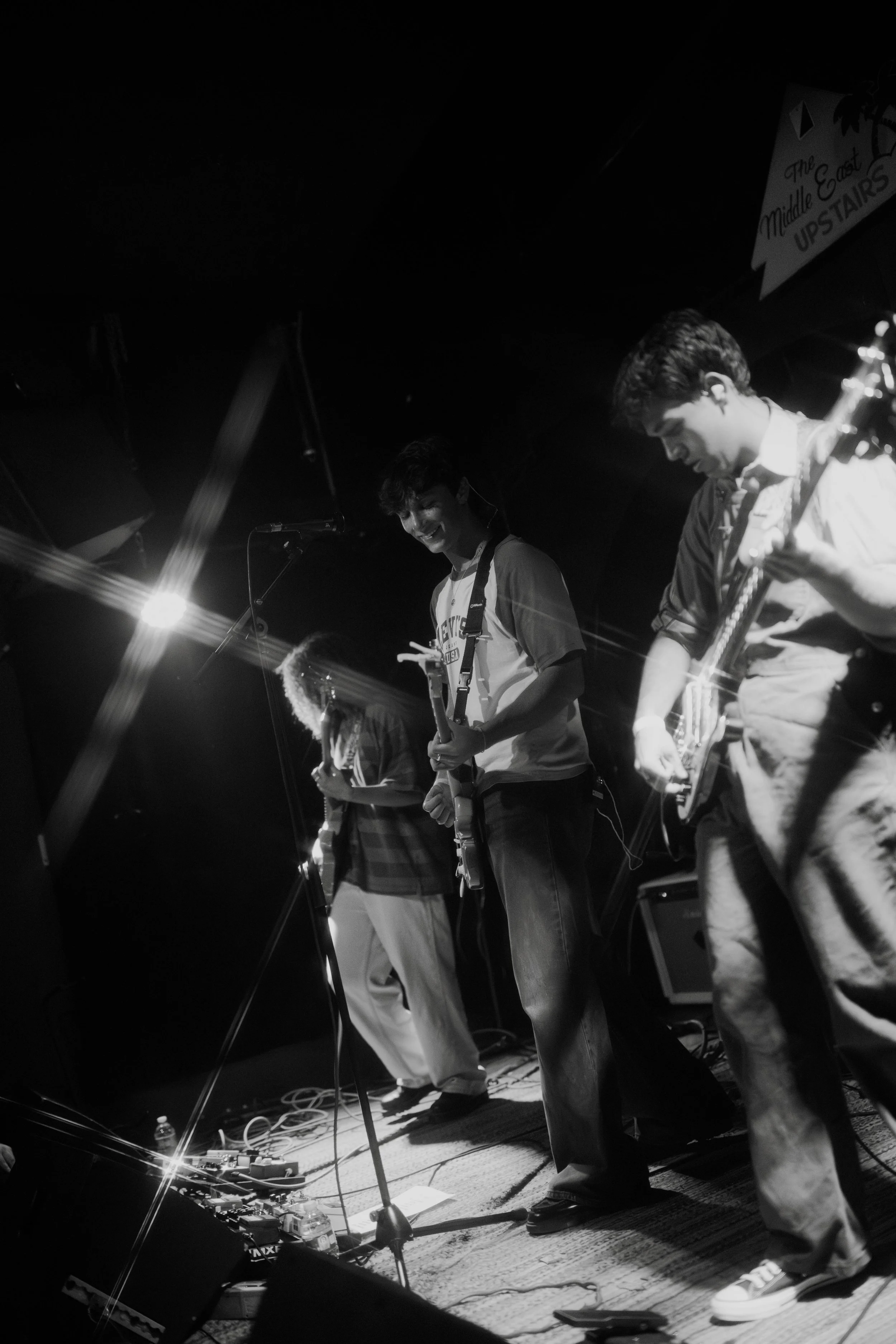 Kevian Kramer and his band performing on stage with three members playing guitars and smiling. There are stage lights and musical equipment around them.