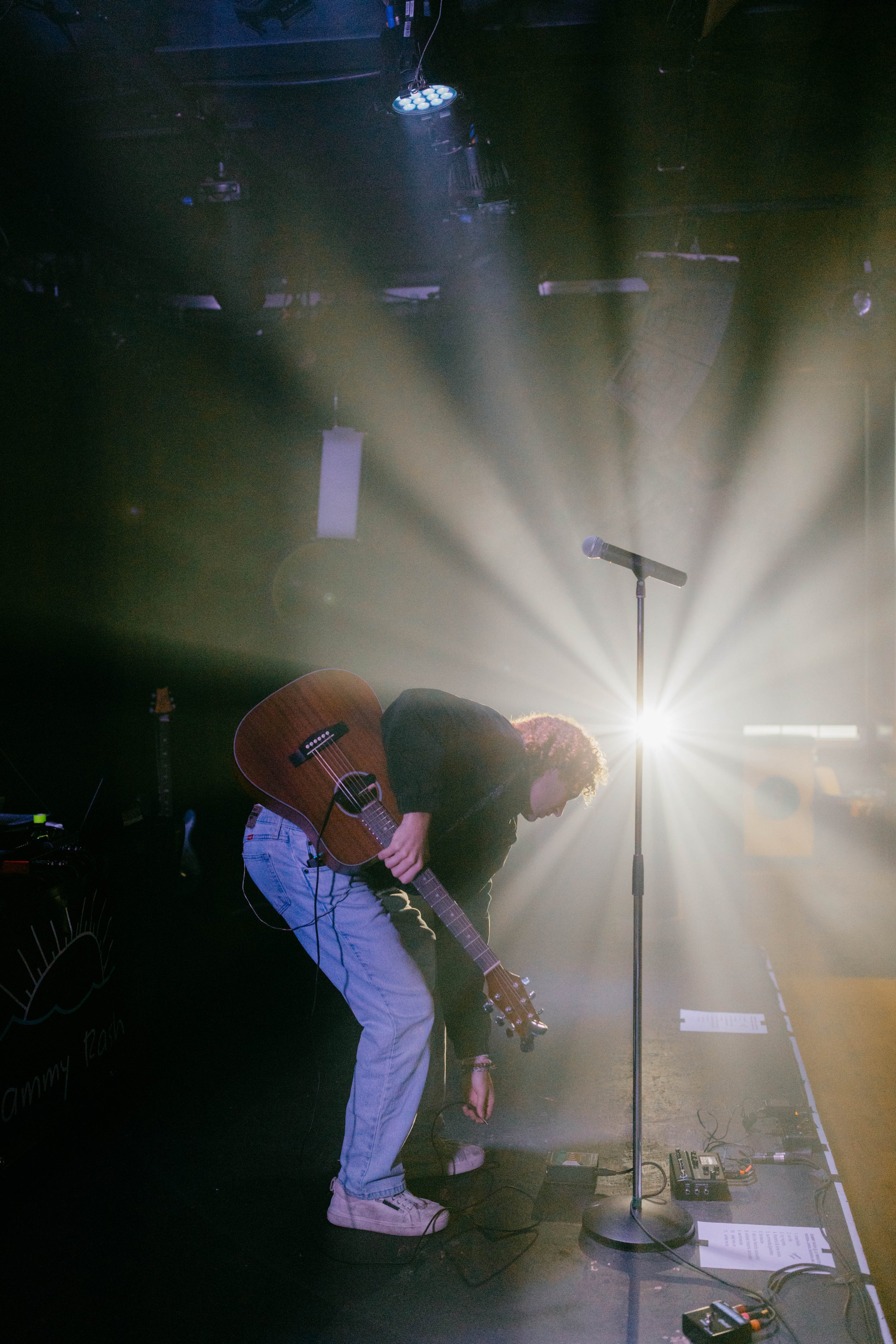 Sammy Rash adjusting equipment on stage with an acoustic guitar on his back, illuminated by bright stage lights creating a starburst effect.