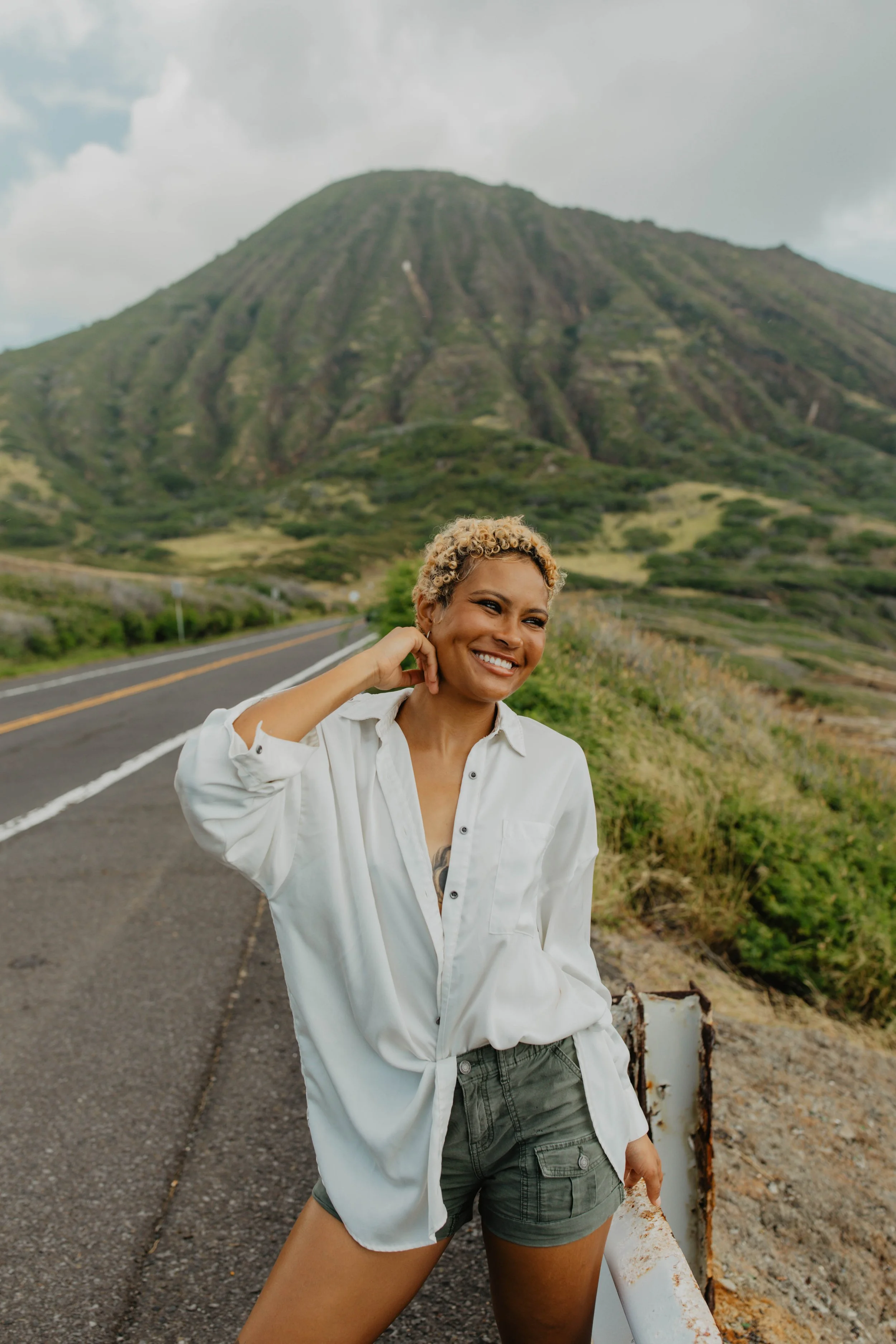 A woman with short blonde curly hair smiling and posing beside a road with a large mountain in the background.