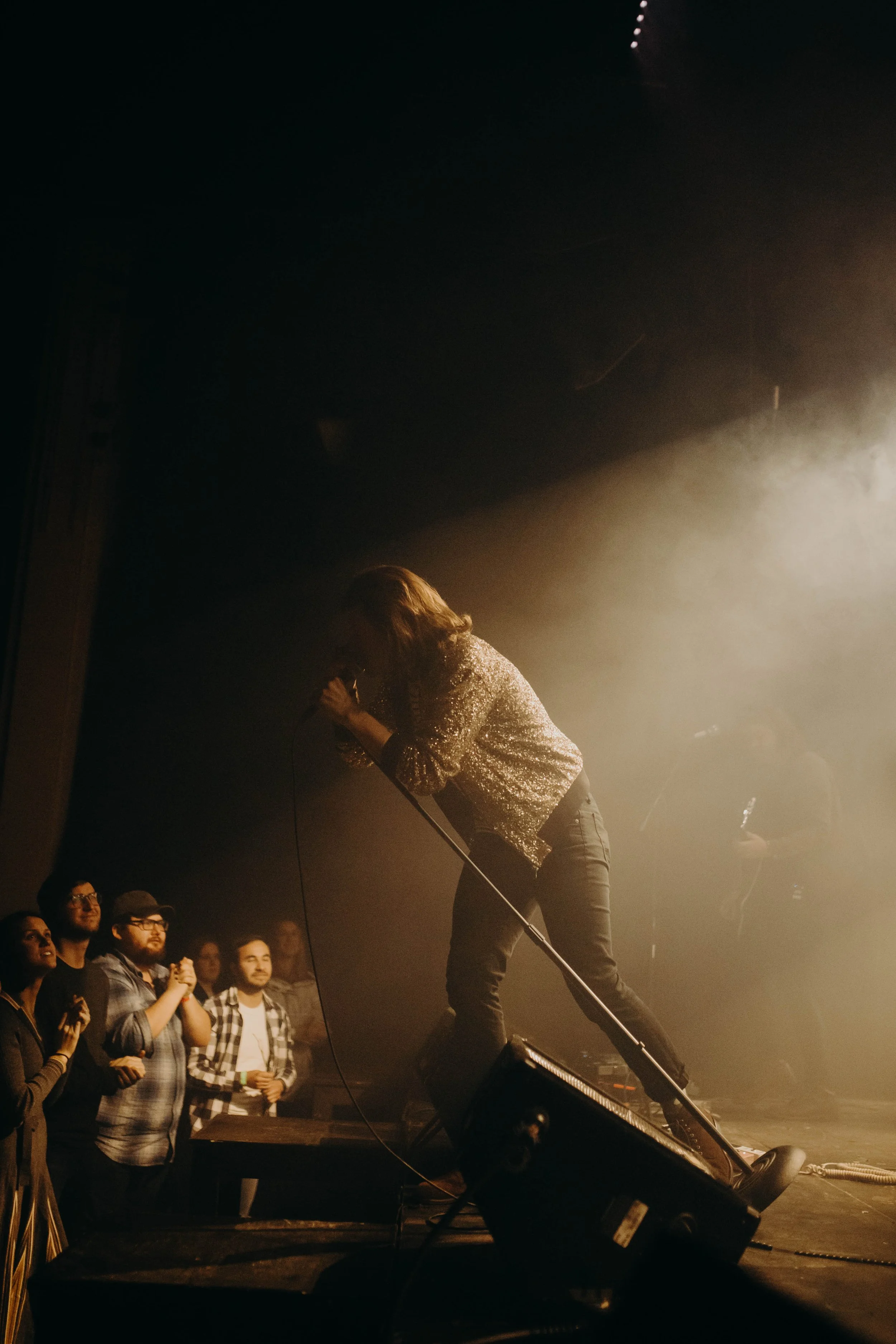 Mike Mains performing on stage with a microphone, surrounded by stage lights and an audience watching and clapping.