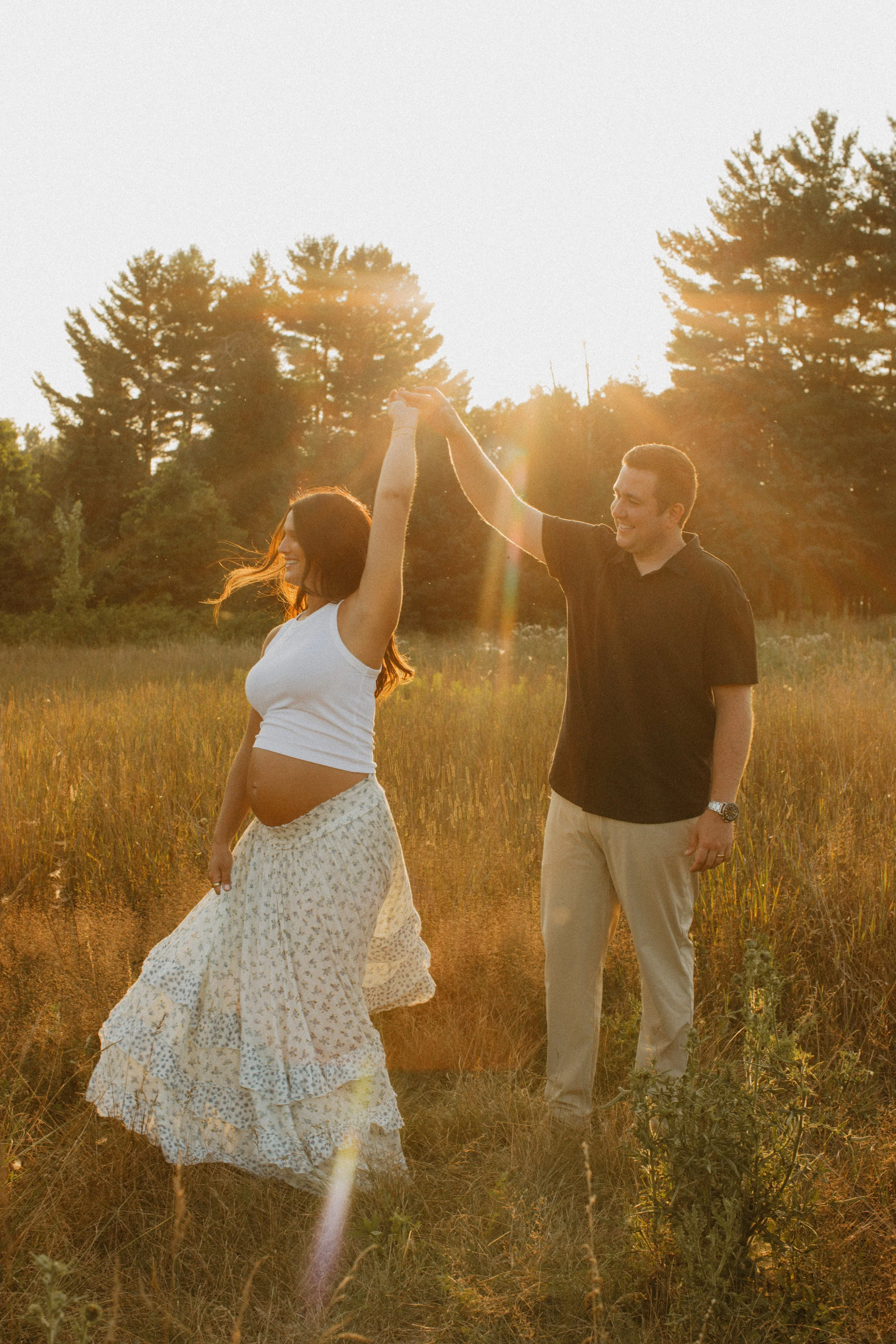 A pregnant woman and a man dancing in a sunlit field, with the woman spinning as the man holds her hand, during sunset.