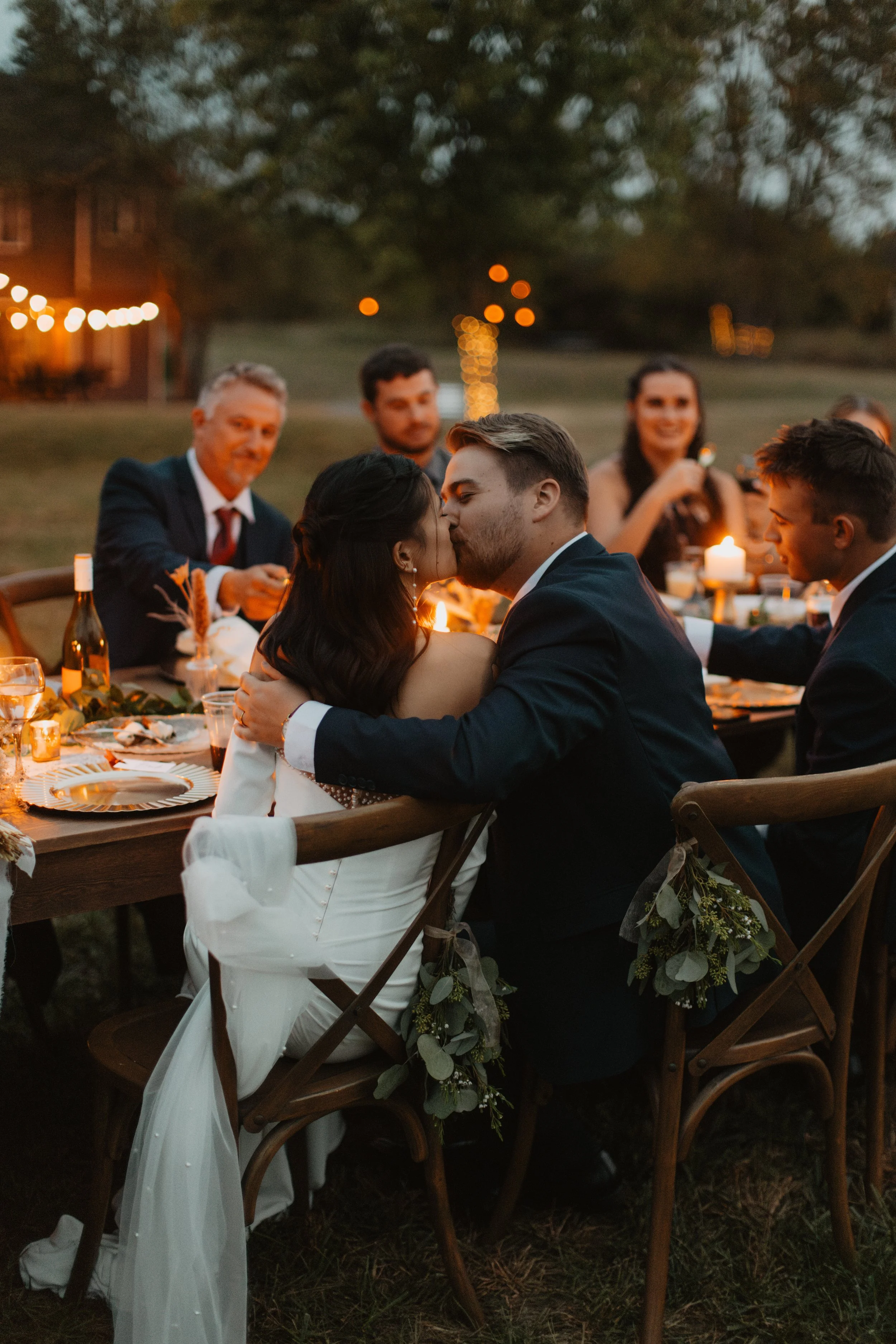 A newlywed couple sharing a kiss at an outdoor wedding reception during the evening, surrounded by friends and family, with warm string lights and candles creating a cozy atmosphere.
