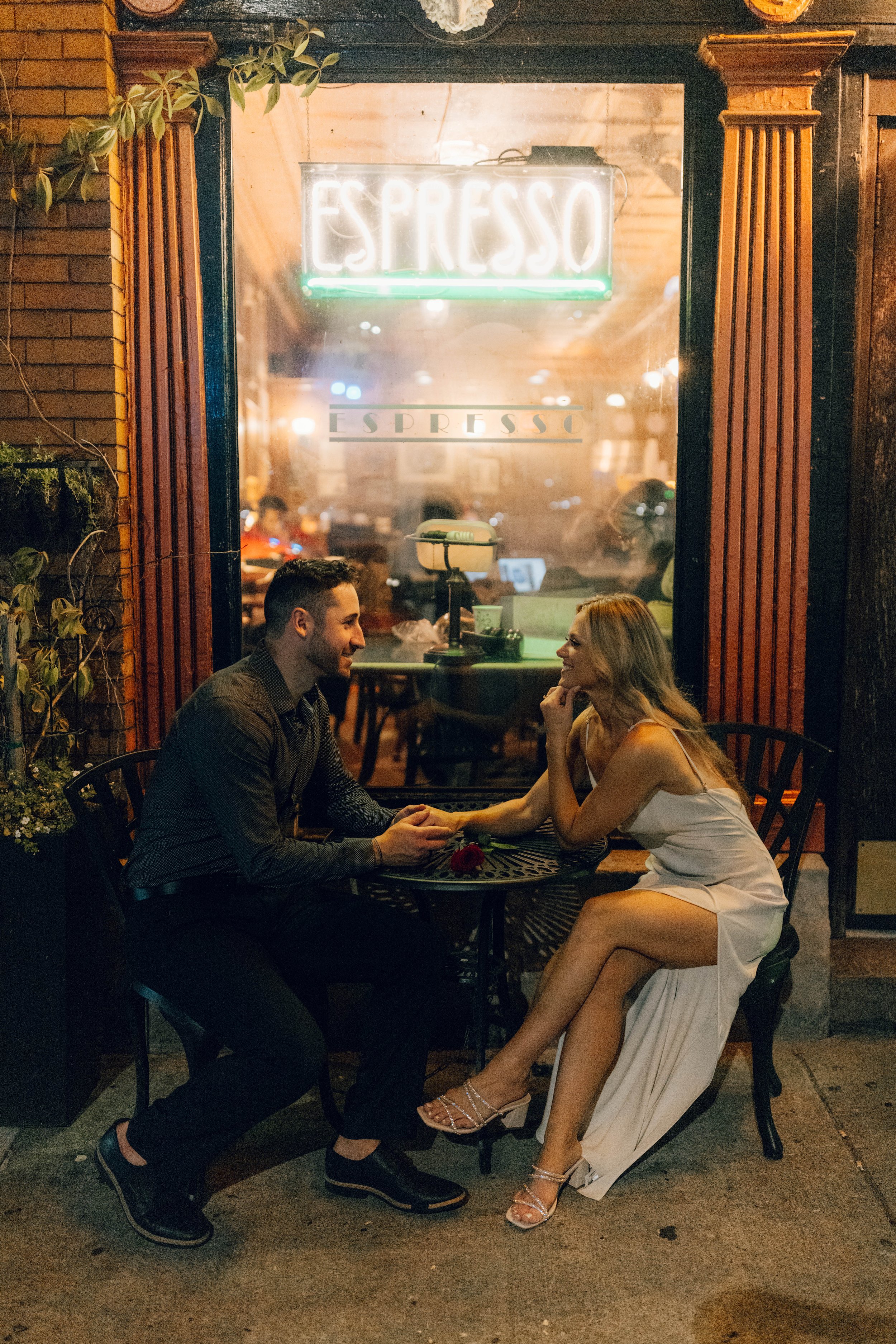 A man and woman are sitting outside a coffee shop at a small round table, holding hands and smiling at each other during evening time. The shop has a neon sign that reads "Espresso" in the window.