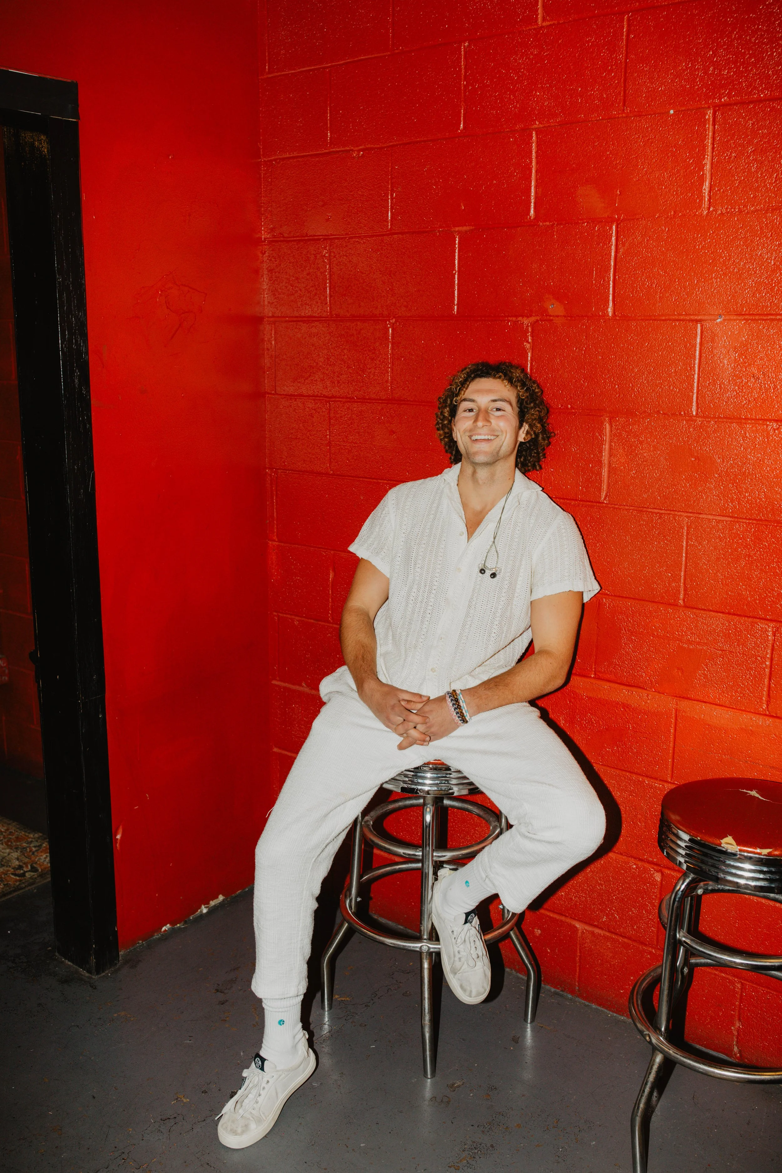 A behind the scenes photo of music artist Sammy Rash smiling while sitting on a stool against a red brick wall, dressed in white pants and a white short-sleeve shirt.