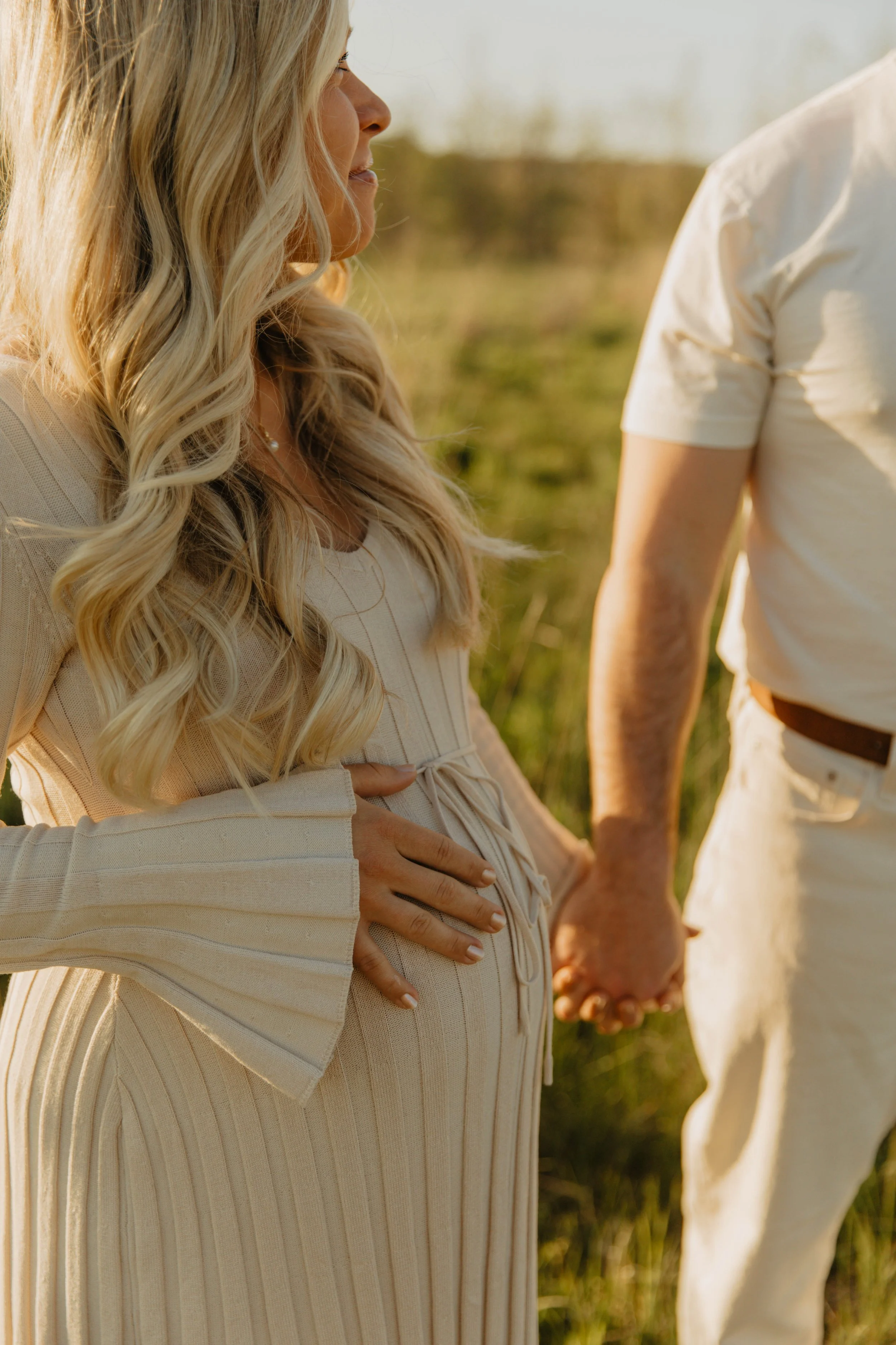 A pregnant woman with long blonde hair holding her belly, standing outdoors while holding hands with a man whose face is not visible.