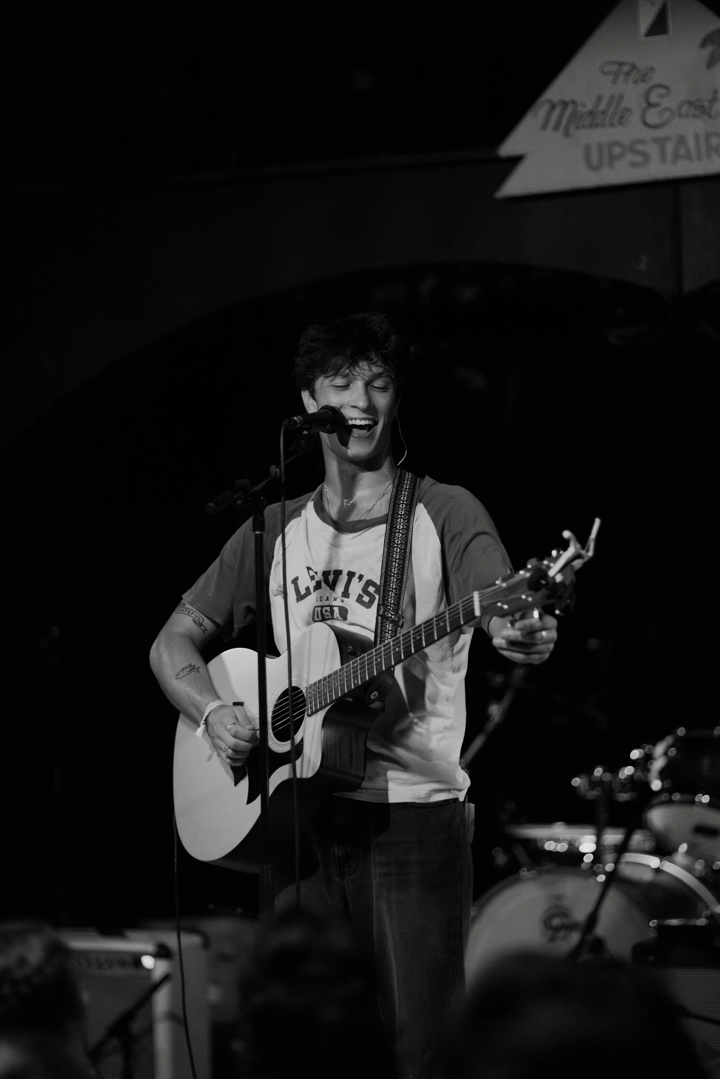 The music artist Kevian Kramer singing into a microphone while playing an acoustic guitar on stage, smiling and wearing a casual t-shirt in Boston.