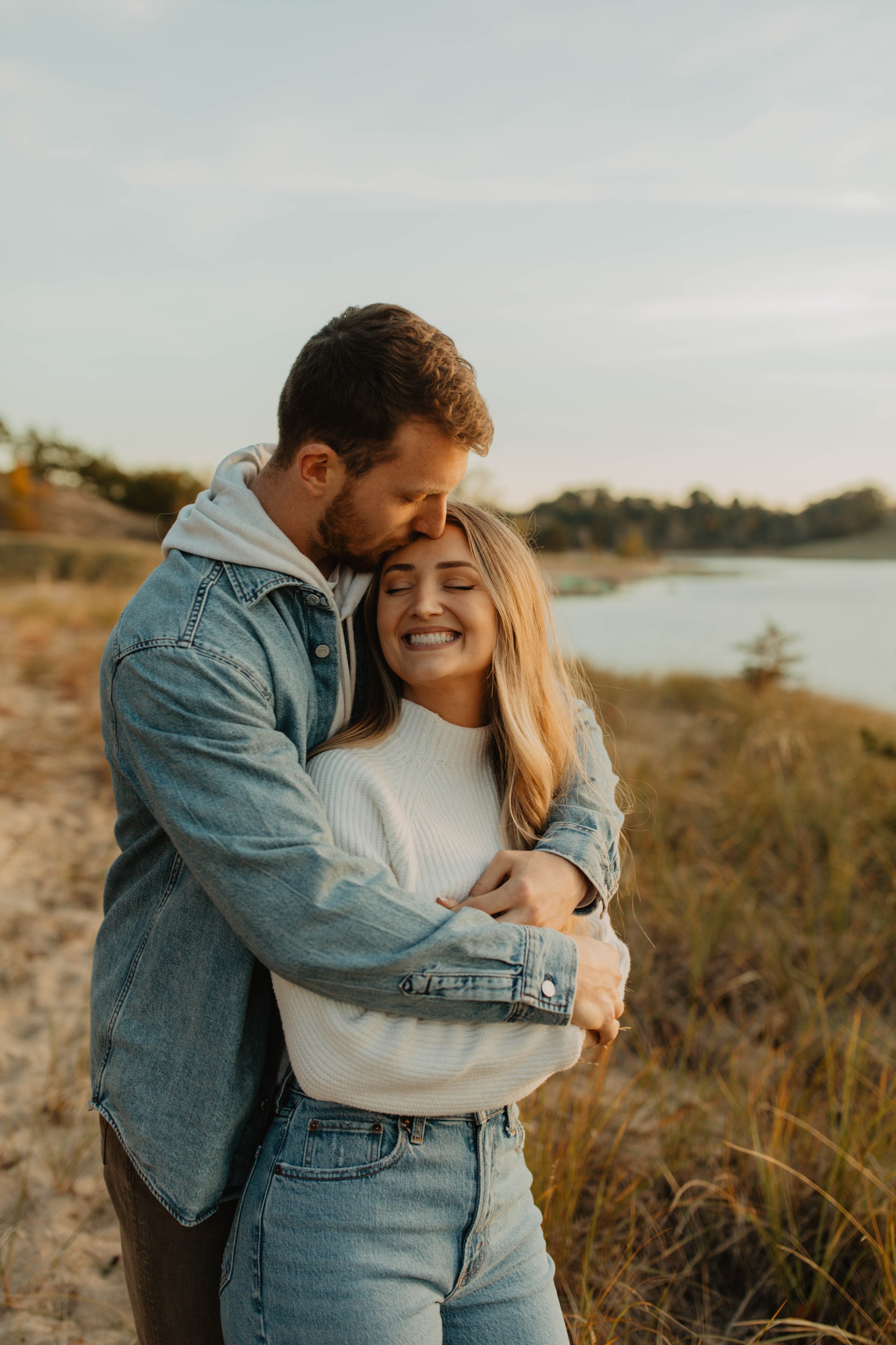 A couple embracing near a lake during sunset, smiling and showing affection.