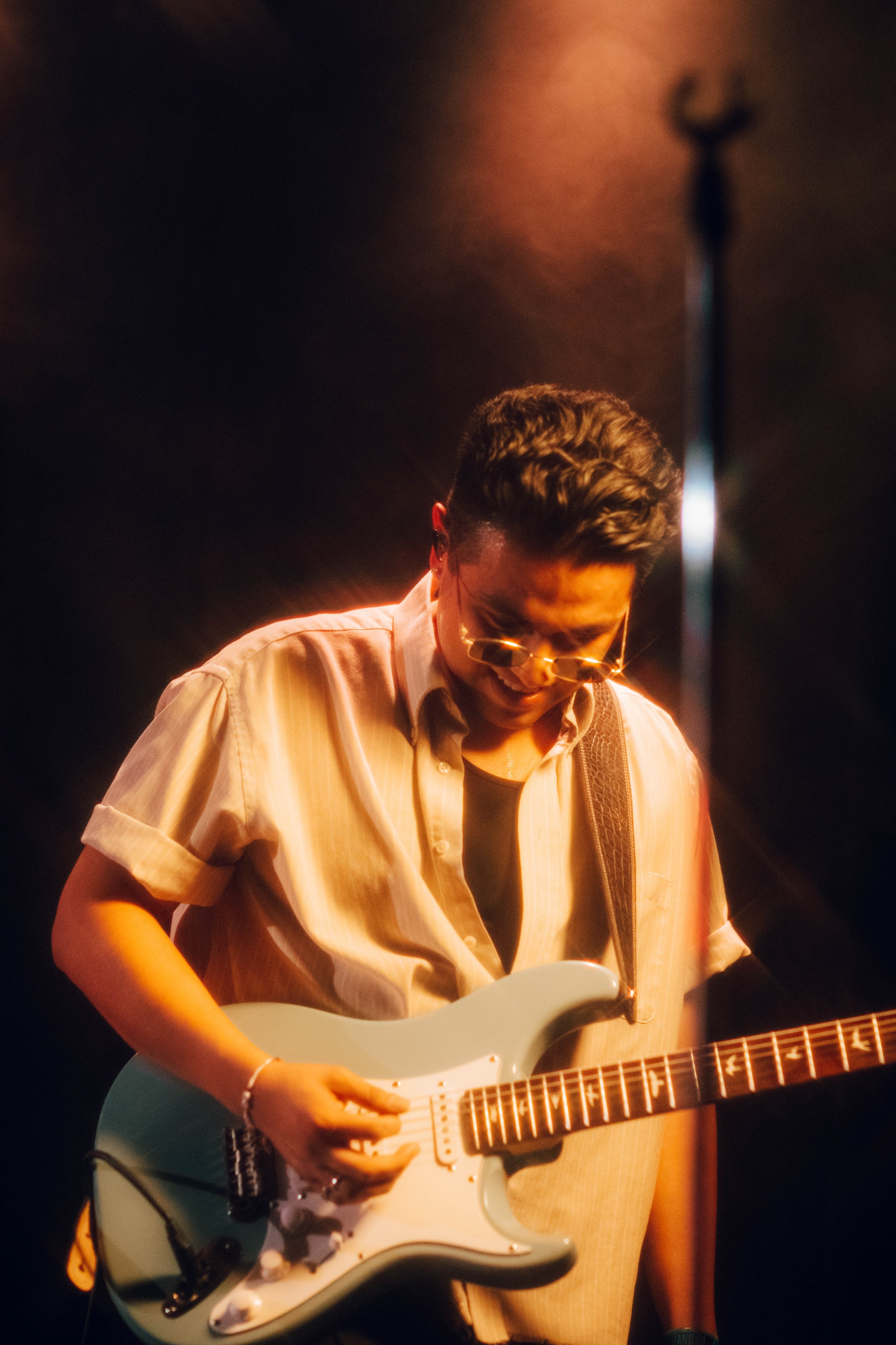 Nicky Youre's band member playing an electric guitar on stage, illuminated by warm stage lighting, with a dark background and a microphone stand nearby.
