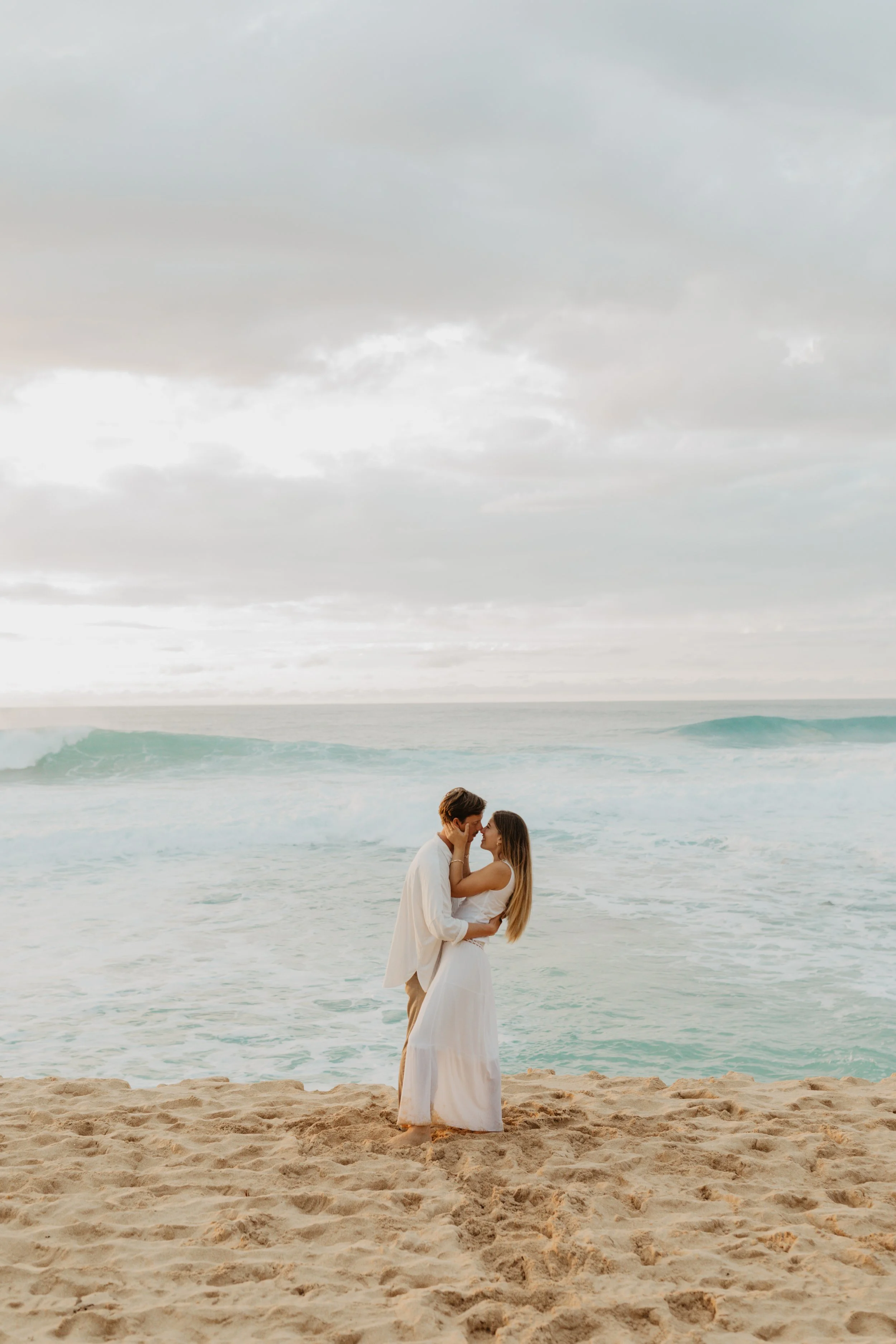A couple embracing on the beach with the ocean and cloudy sky in the background.