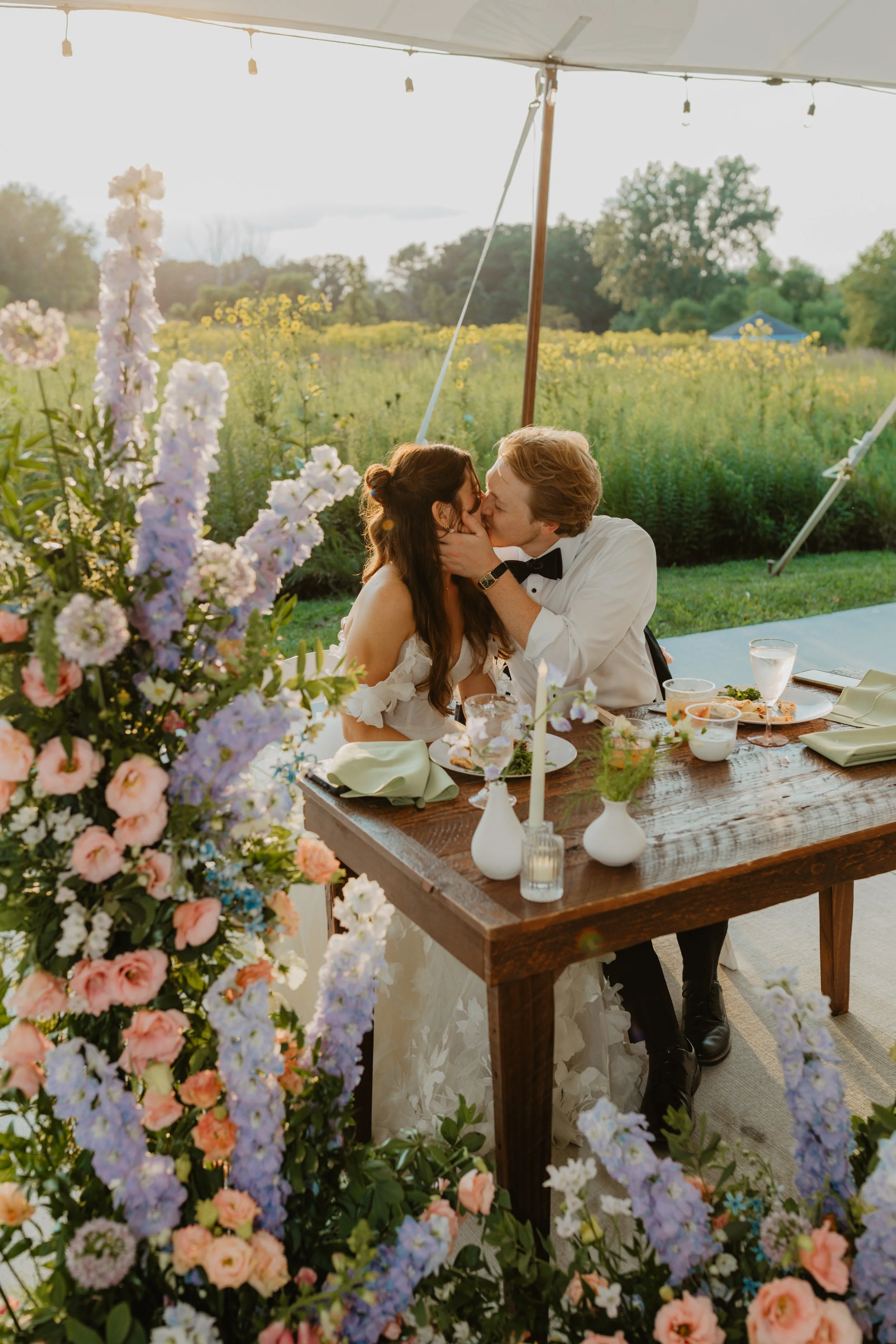 A couple in wedding attire sharing a kiss at a wedding reception outdoors, with flowers and a field in the background.