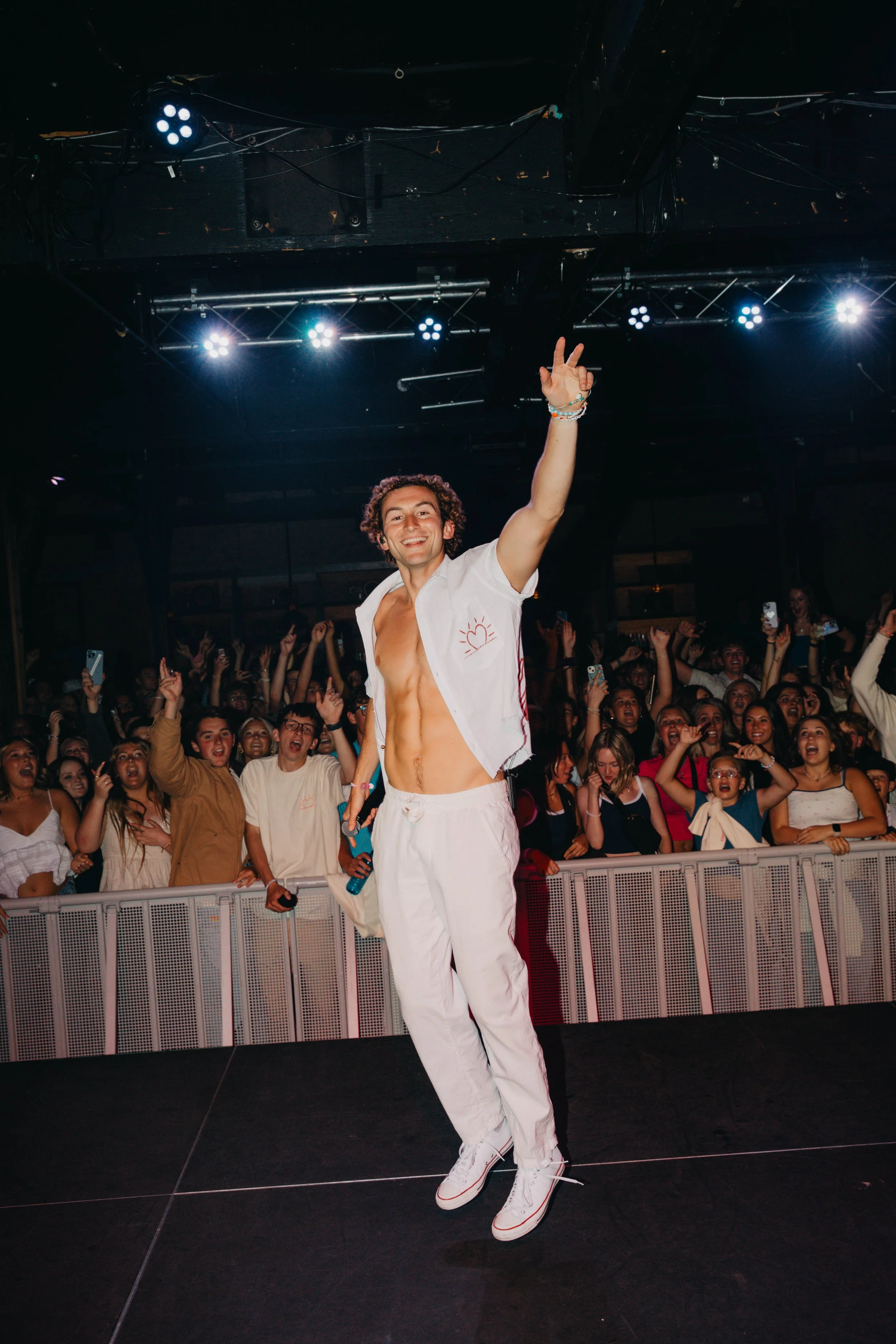 A young man with curly hair, shirt open to reveal his torso, dancing on stage during a concert, with an excited crowd in the background.