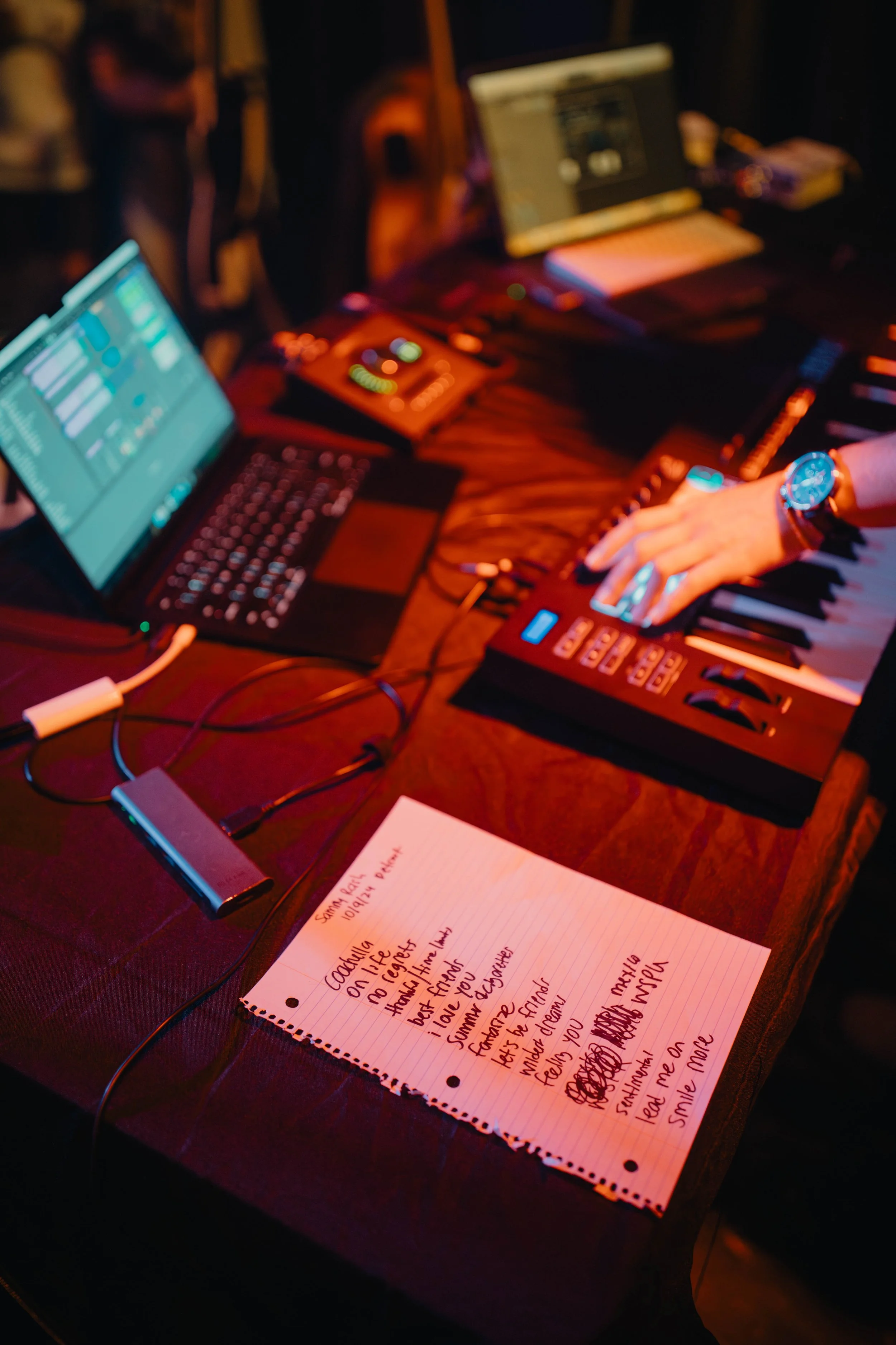 A music production setup on a wooden table with a laptop, a MIDI controller, a notebook with handwritten lyrics, and a person's hand playing the keyboard. The scene is illuminated with orange and pink lighting.