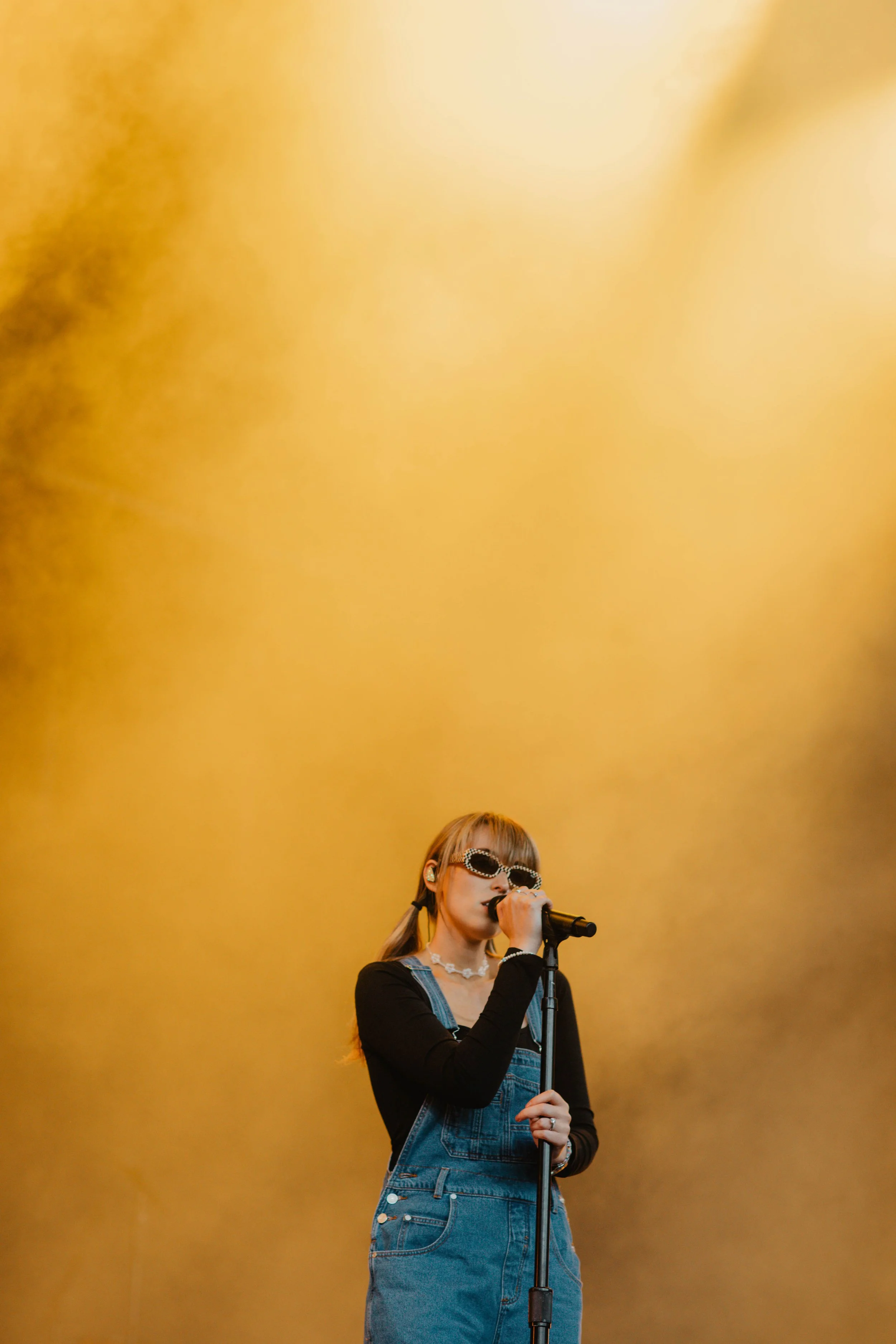 A music artist Sasha Sloan singing into a microphone on stage, wearing sunglasses, a black long-sleeve shirt, and denim overalls, with a warm yellow background.