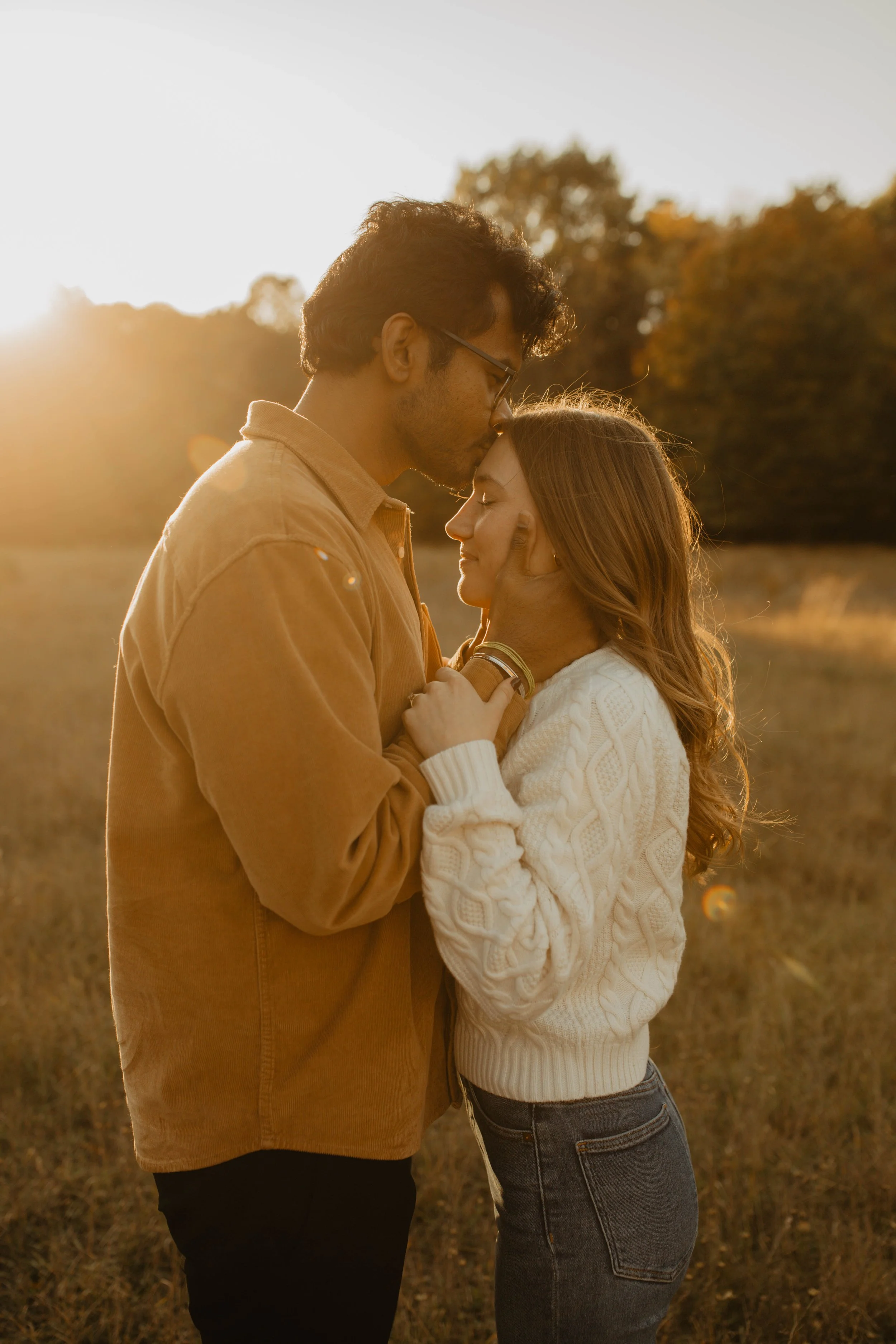 A couple sharing an intimate moment outdoors at sunset, with the man kissing the woman's forehead as they hold each other.