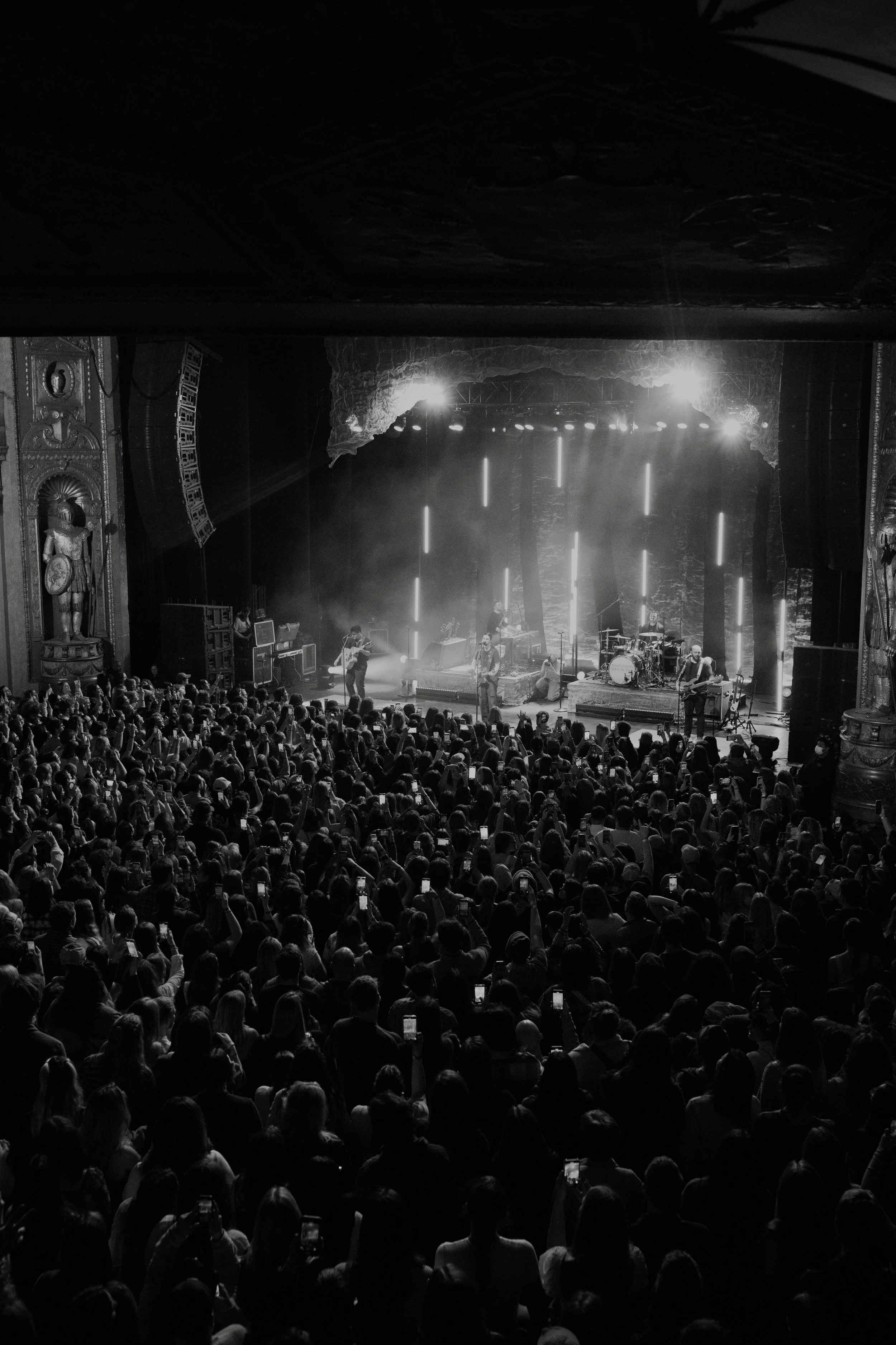 A black and white photo of a live concert with a large audience, stage lighting, and a band performing.