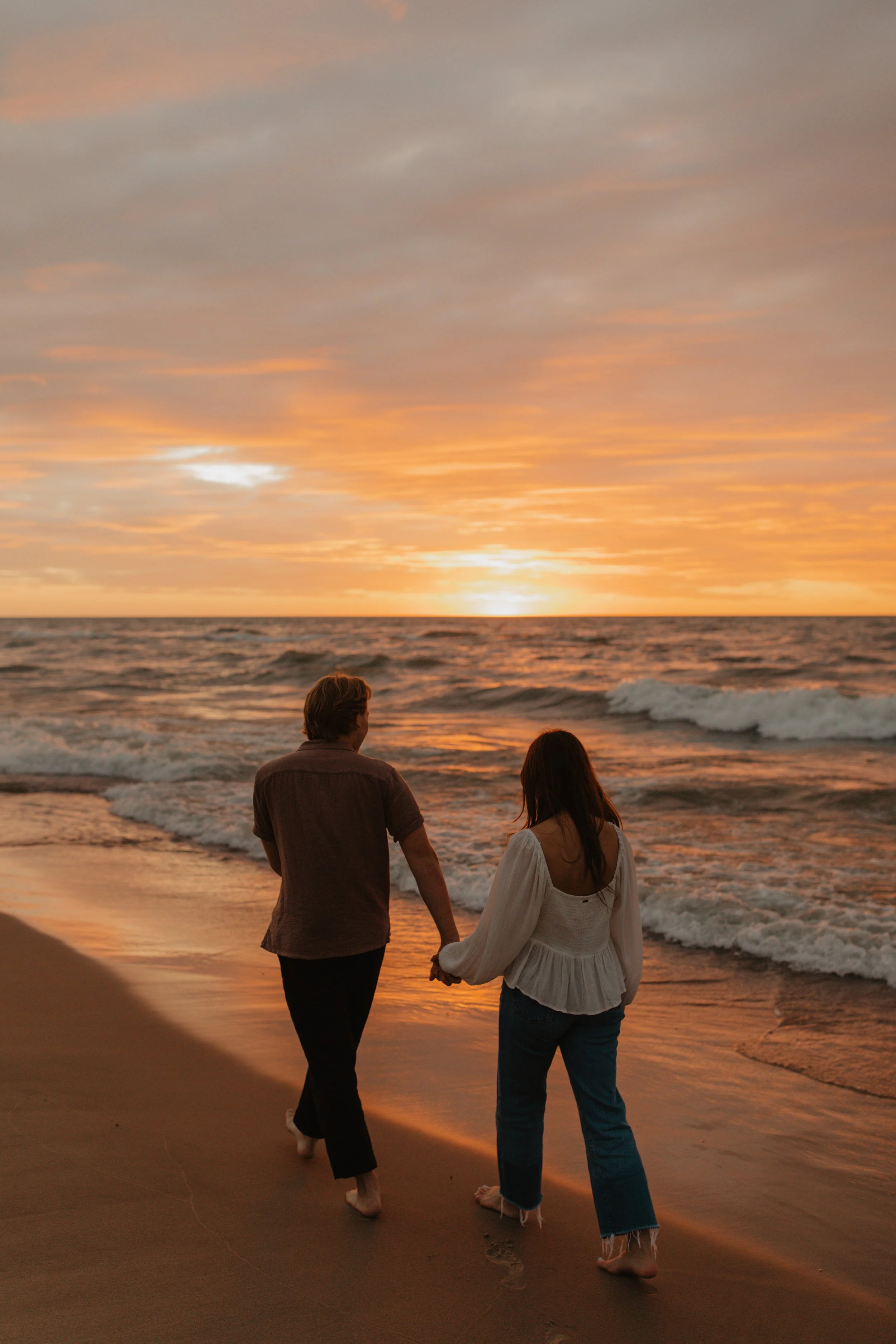 A man and woman walking hand in hand on the beach during sunset.