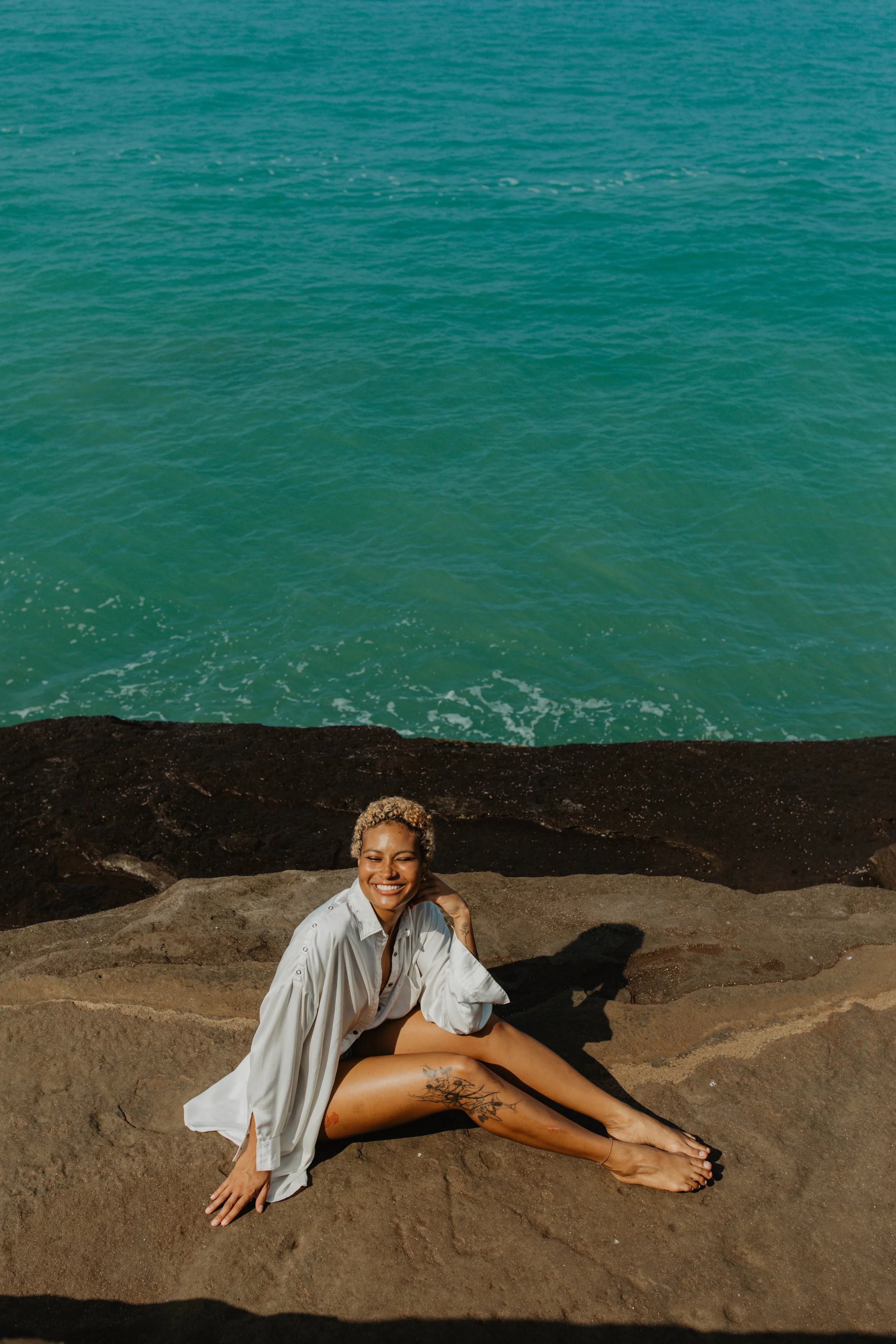 A woman with short curly blonde hair and tattoos, smiling and sitting on rocks near the ocean on a sunny day.